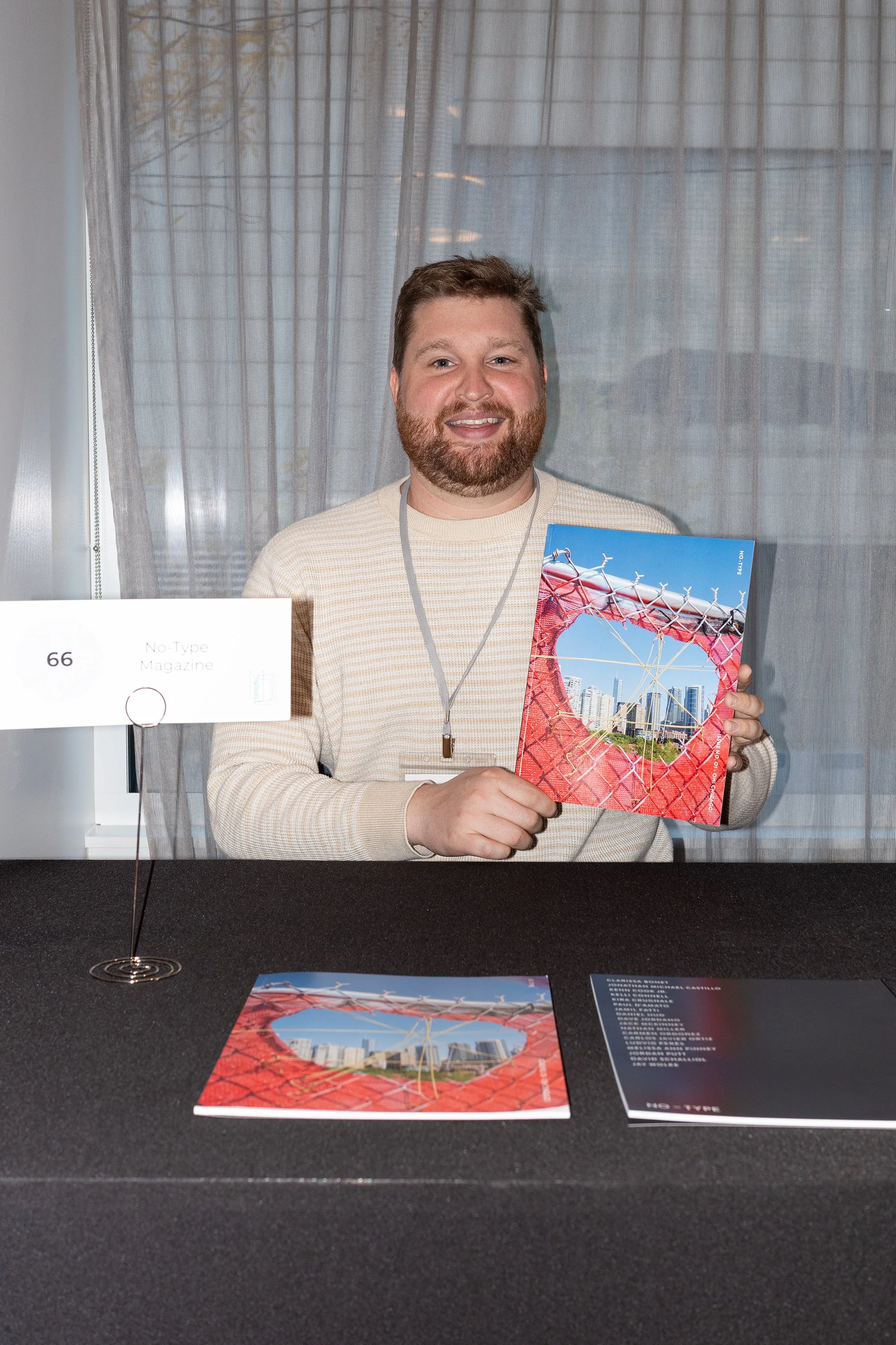 A man with a beard and light brown hair, wearing a beige sweater, sitting behind a table at a convention or event, holding a colorful magazine featuring a cityscape through a red, circular, geometric structure. There is a sign with the number 66 and the text "No-Type Magazine" on the table, along with another copy of the magazine and some literature or flyers.