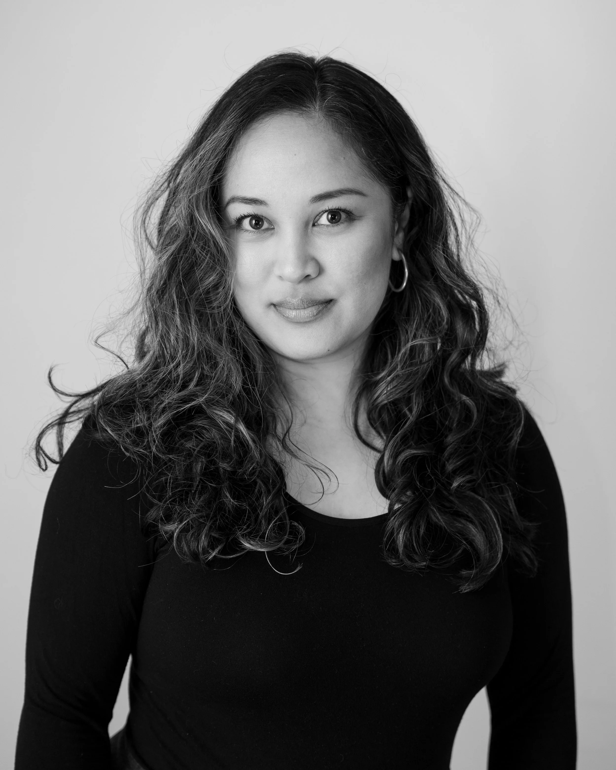 Black and white portrait of a woman with wavy hair wearing a black top and hoop earrings, looking at the camera.