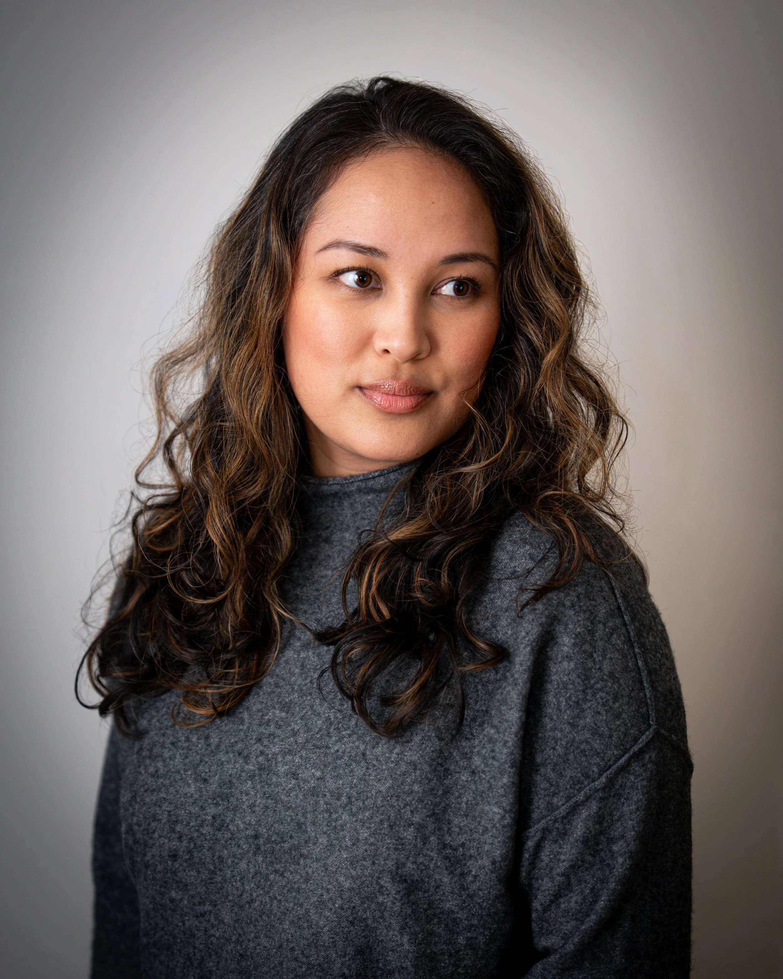 Portrait of a woman with wavy brown hair wearing a dark gray sweater, looking slightly to the side, against a plain neutral background.