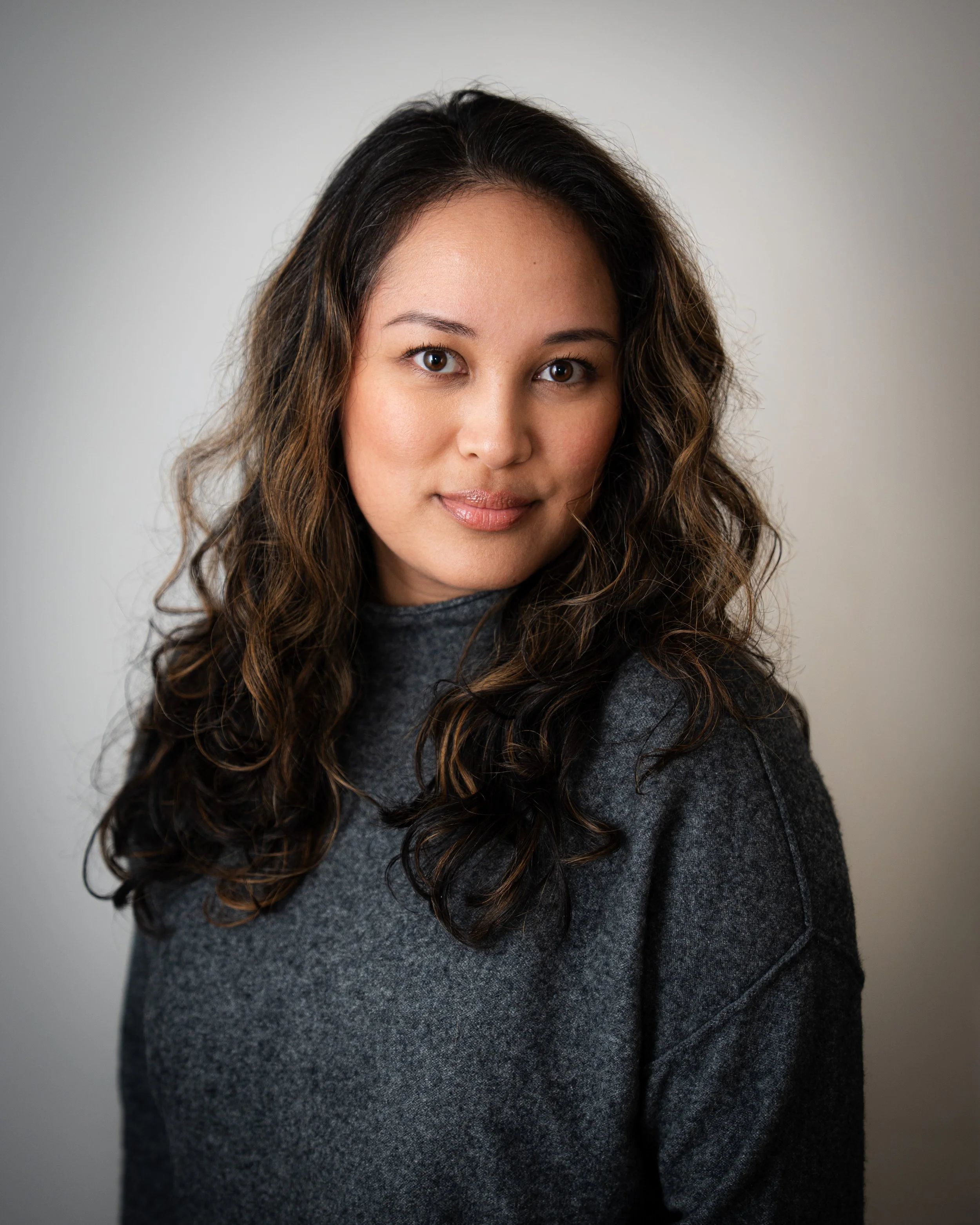 Portrait of a woman with long, wavy dark hair, wearing a gray sweater, smiling softly against a plain light background.