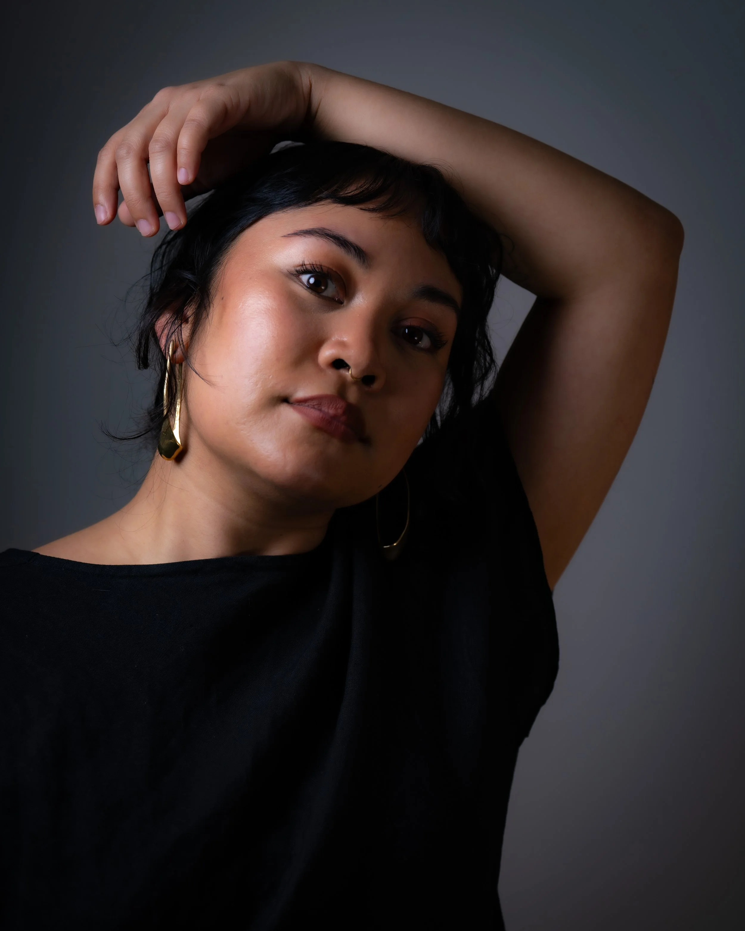 Portrait of a woman with short dark hair, wearing a black top, gold earrings, and a septum piercing, posing with one arm raised above her head against a plain dark background.