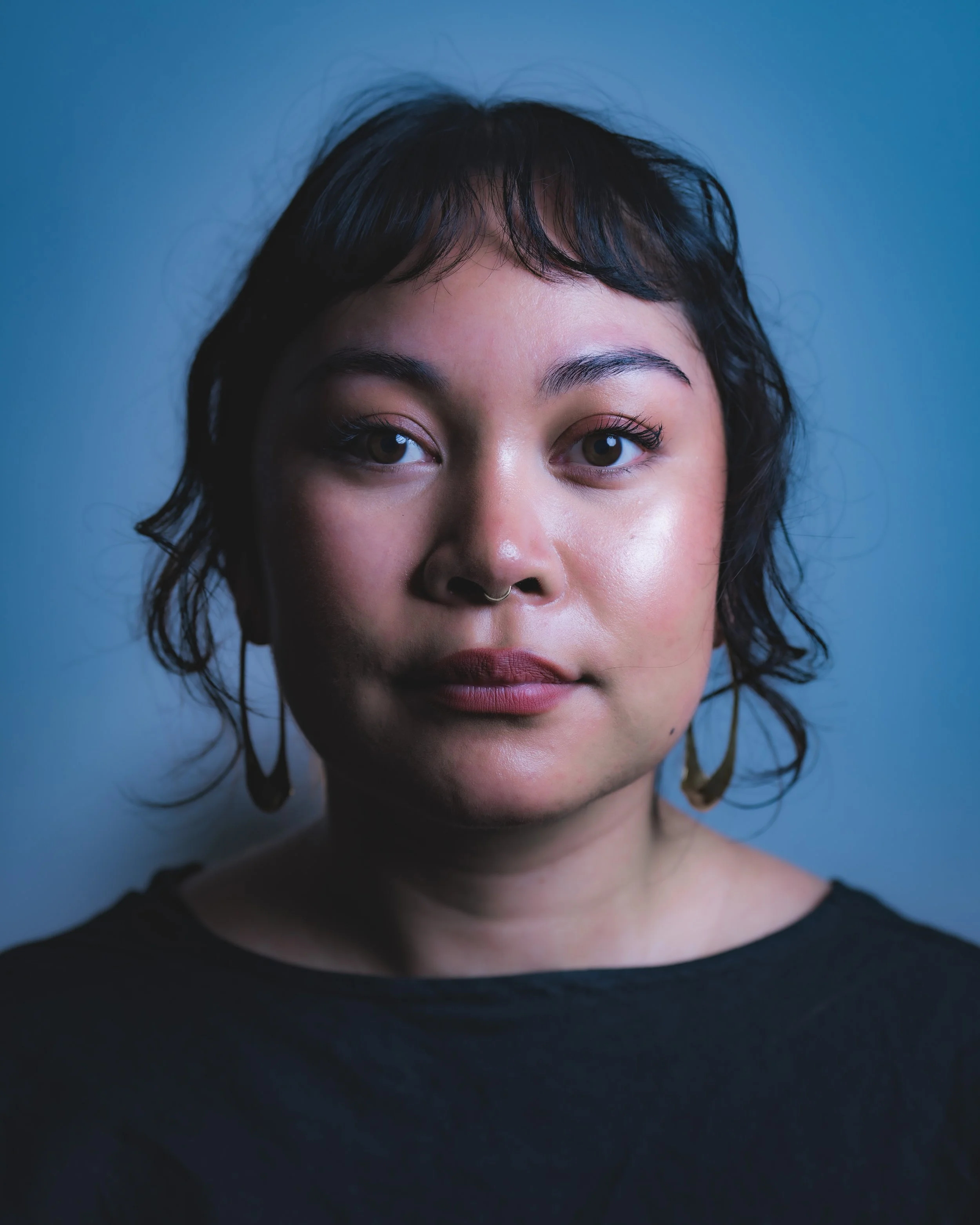 A woman with dark curly hair, wearing earrings and a black top, looking directly at the camera with a neutral expression against a blue background.