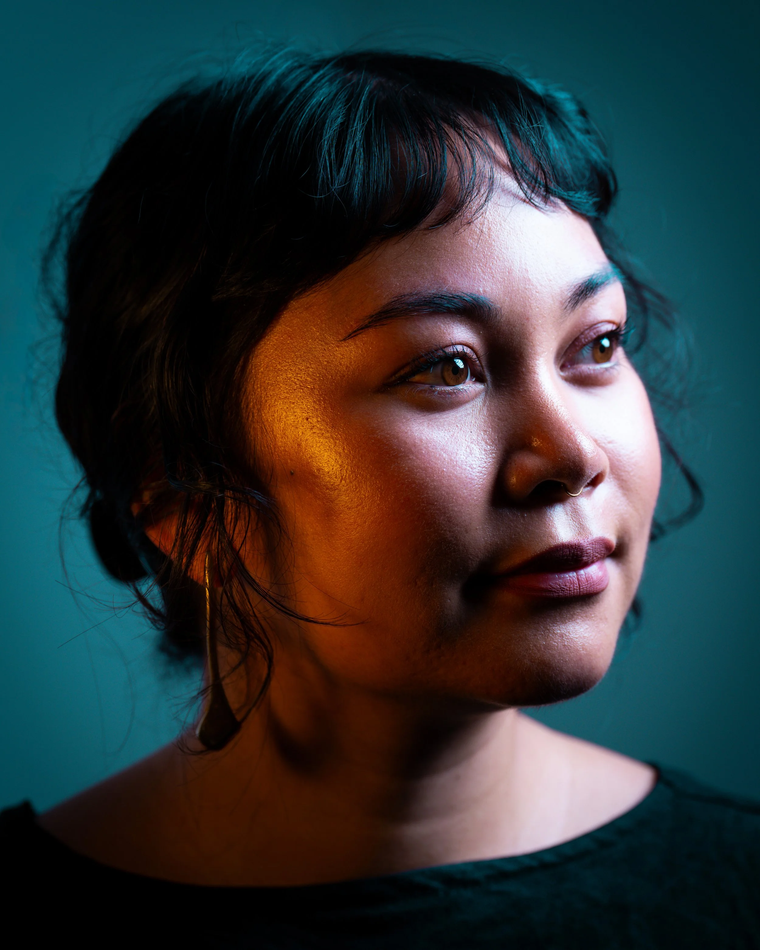 Close-up portrait of a woman with short black hair, brown eyes, wearing hoop earrings, and a septum piercing, against a dark background.