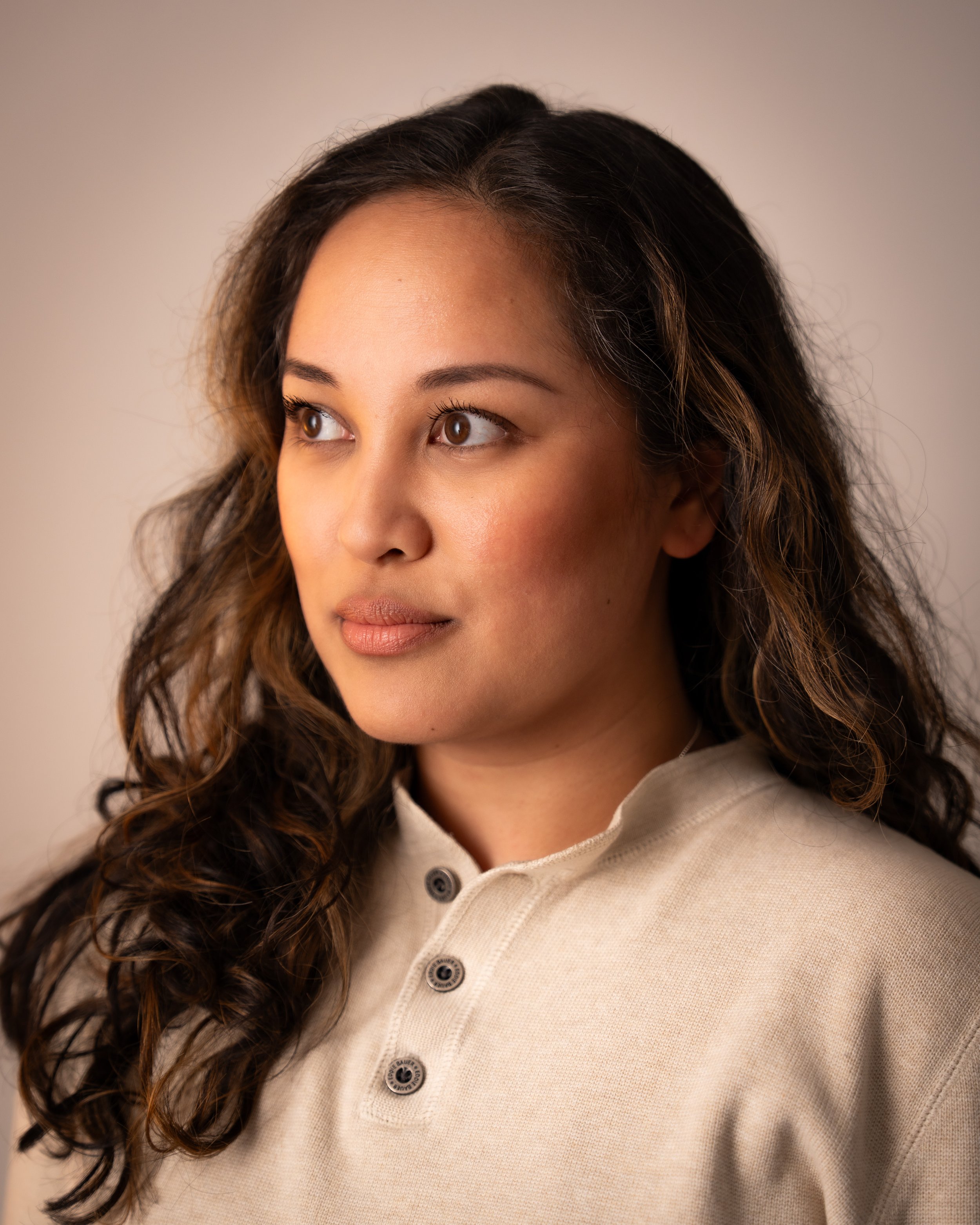 A woman with long, wavy brown hair looking to the side wearing a beige collared shirt with buttons.