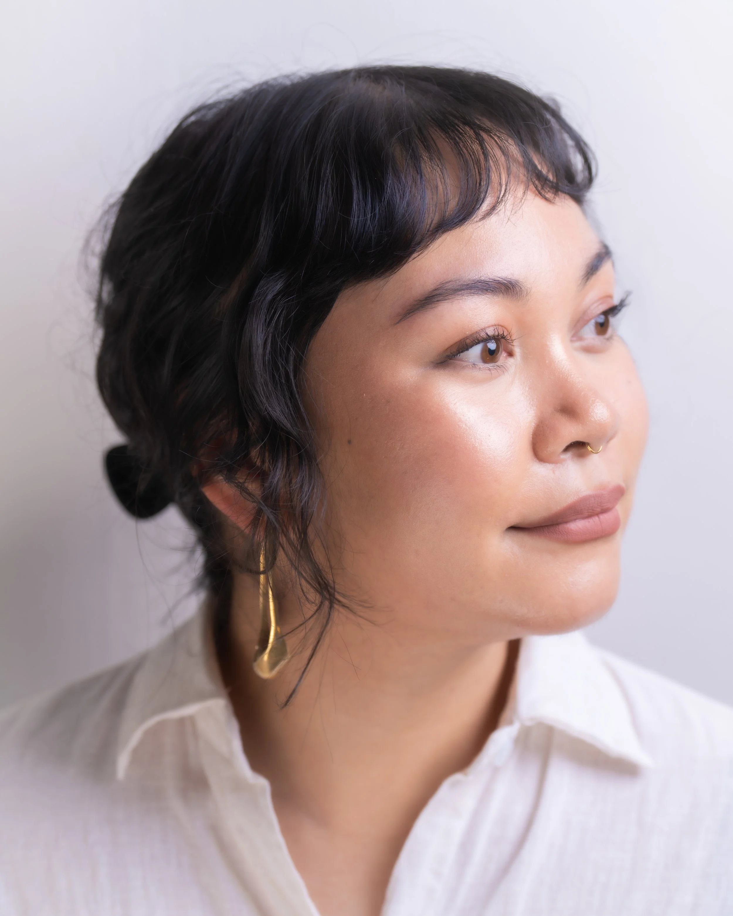 A woman with short, wavy black hair, light skin, wearing gold earrings and a white shirt, looking to her right.