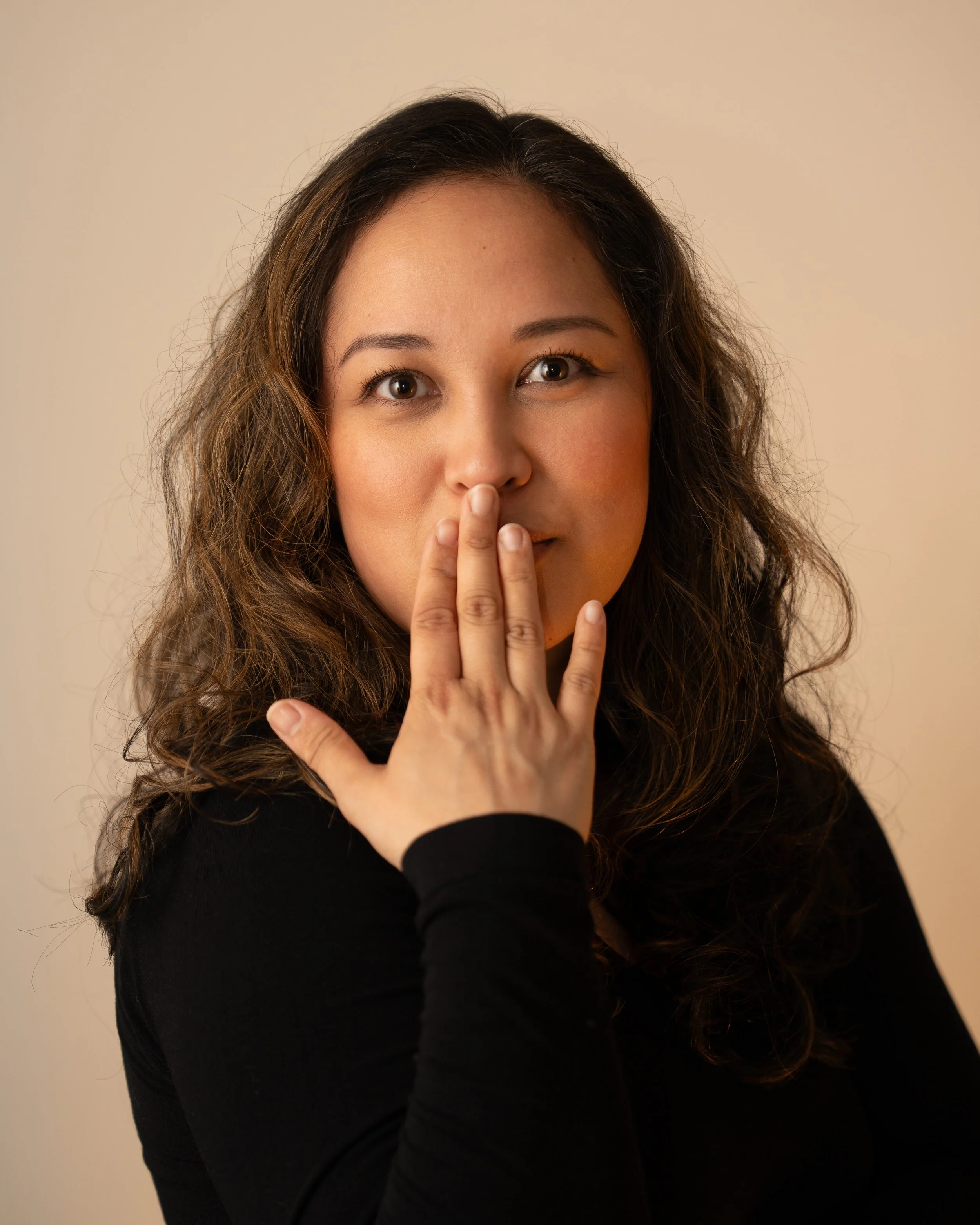 A woman with long, wavy brown hair and a black top is holding her hand to her lips in a hush gesture, looking directly at the camera against a neutral background.