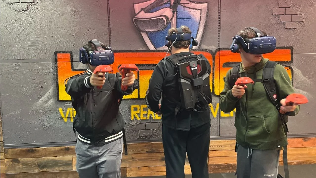 Three players wearing VR headsets, controllers, and backpack equipment during an immersive group VR gaming session.