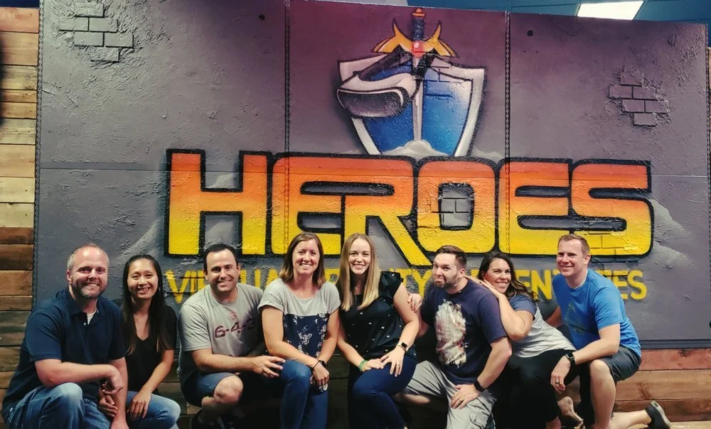 Group of eight people smiling and sitting in front of a large sign that says 'Heroes' with a shield and sword logo, in a casual indoor setting.
