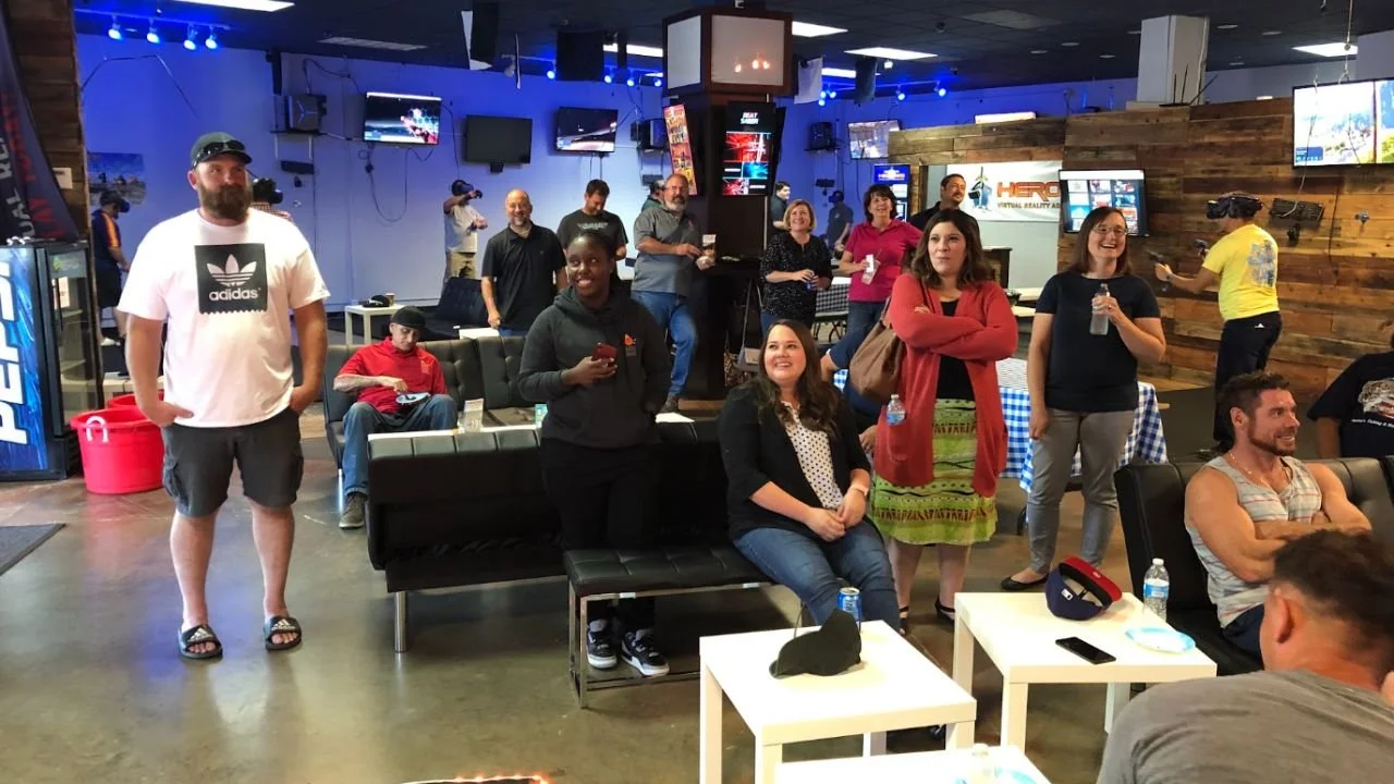 Adults gathering in the lounge at HEROES VR Adventures while players enjoy VR games nearby.