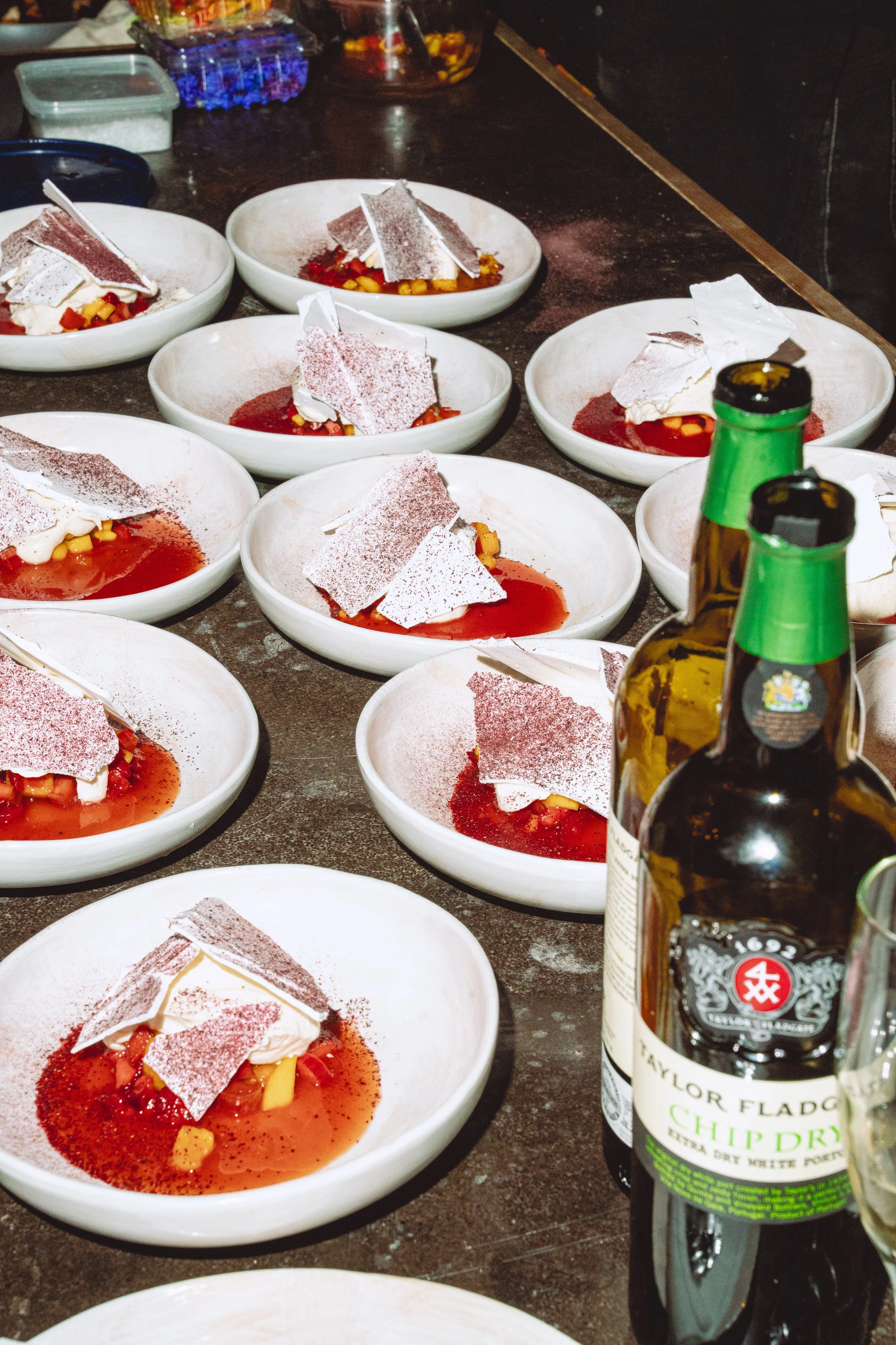 Several bowls with a dessert topped with a slice of white meringue, red sauce, and small yellow fruit pieces, along with beer bottles on a black countertop.