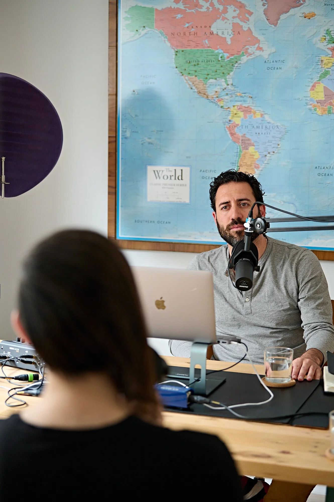 A man with dark curly hair and a beard speaking into a microphone during a podcast or radio recording, with a world map on the wall behind him.