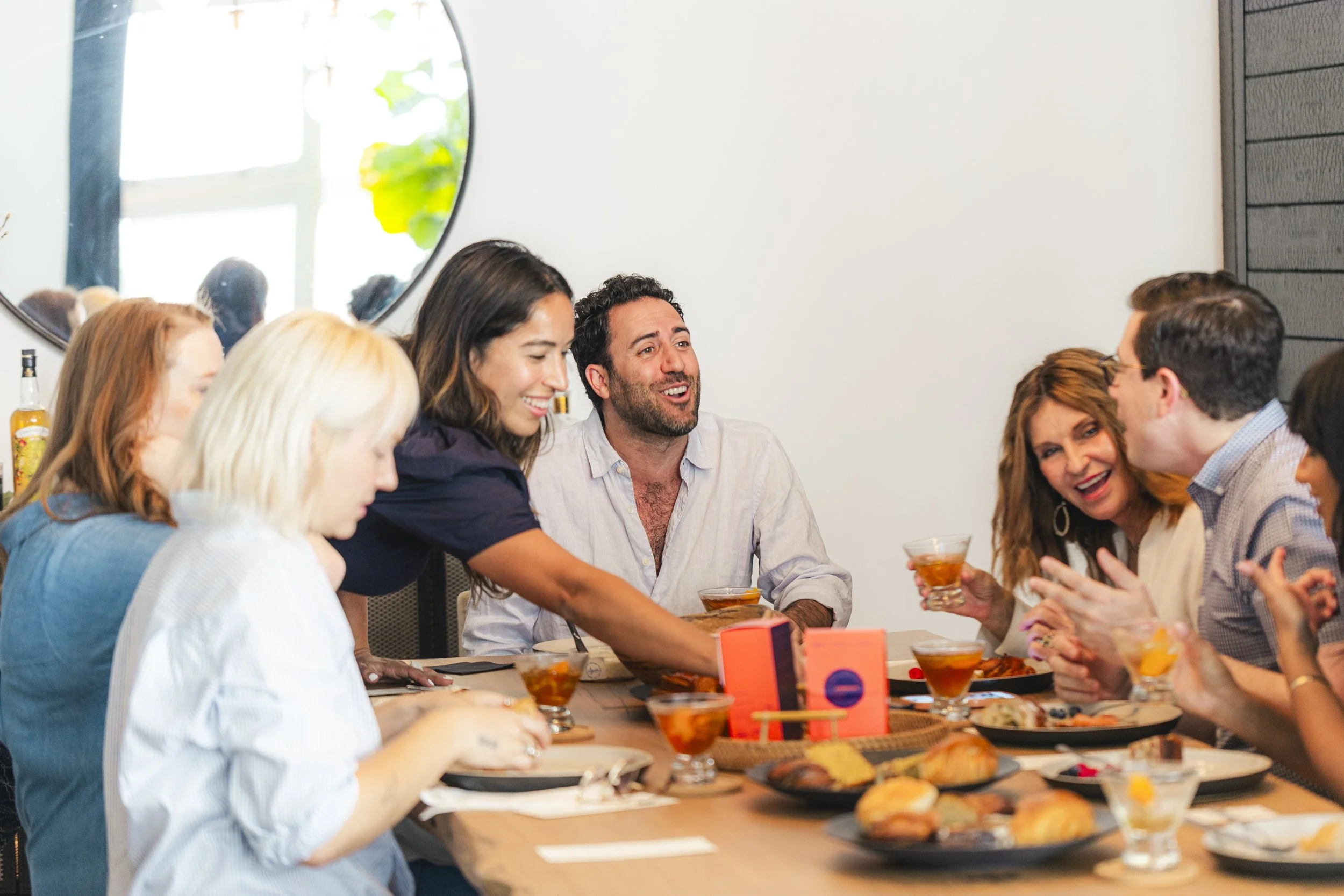 Group of people sitting around a dining table, enjoying a meal and drinks, engaged in conversation, indoors with a large mirror and modern decor.