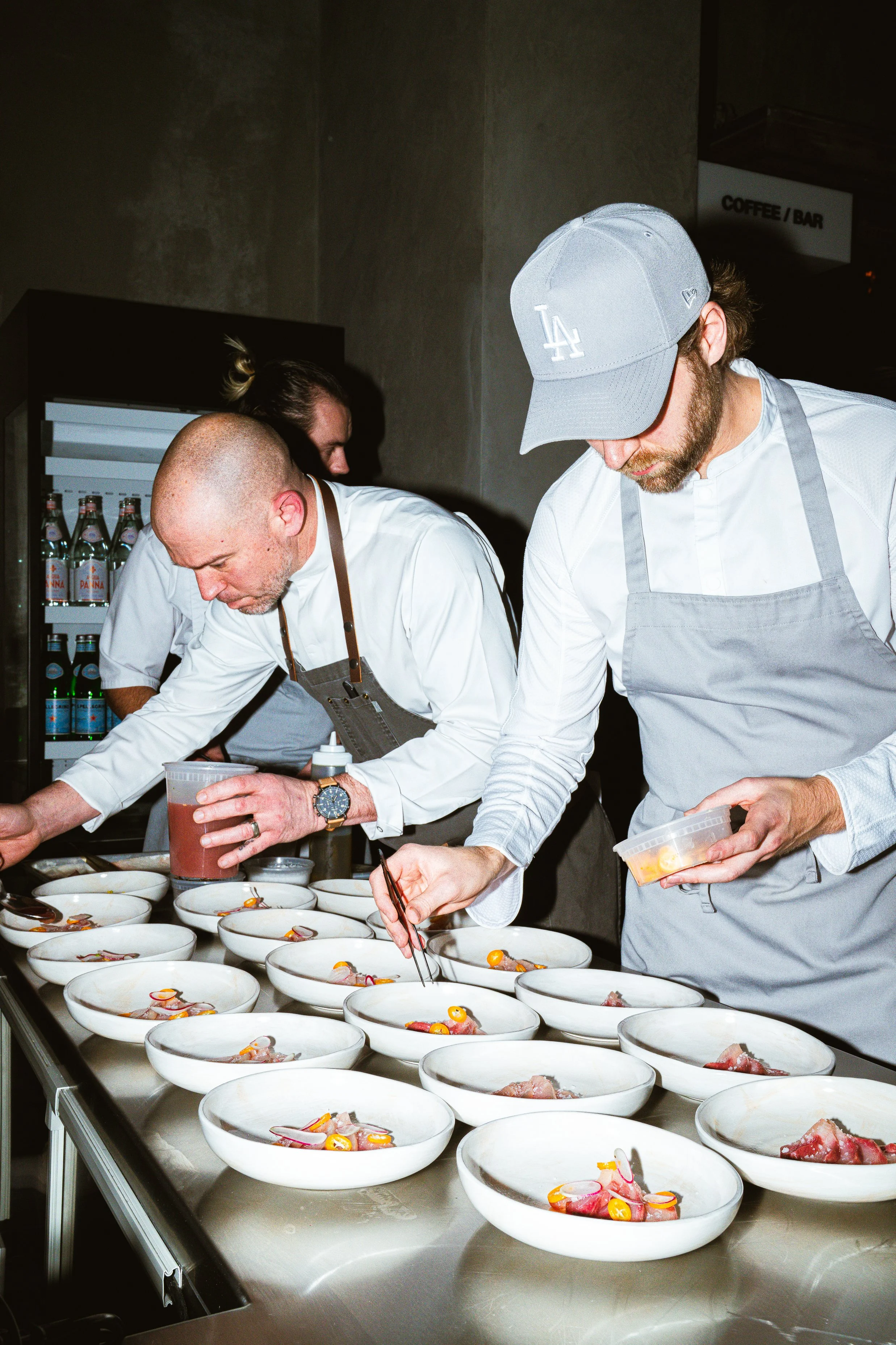 Chefs preparing plated dishes in a professional kitchen, with bowls of food arranged on a stainless steel counter.