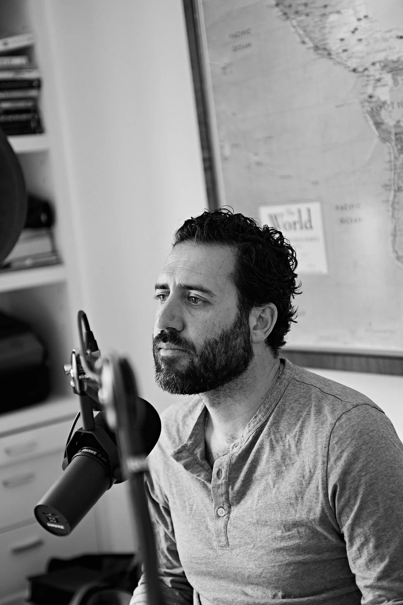 A man with curly hair and a beard sitting at a desk, looking at a computer screen, speaking into a microphone, in a room with a world map on the wall.