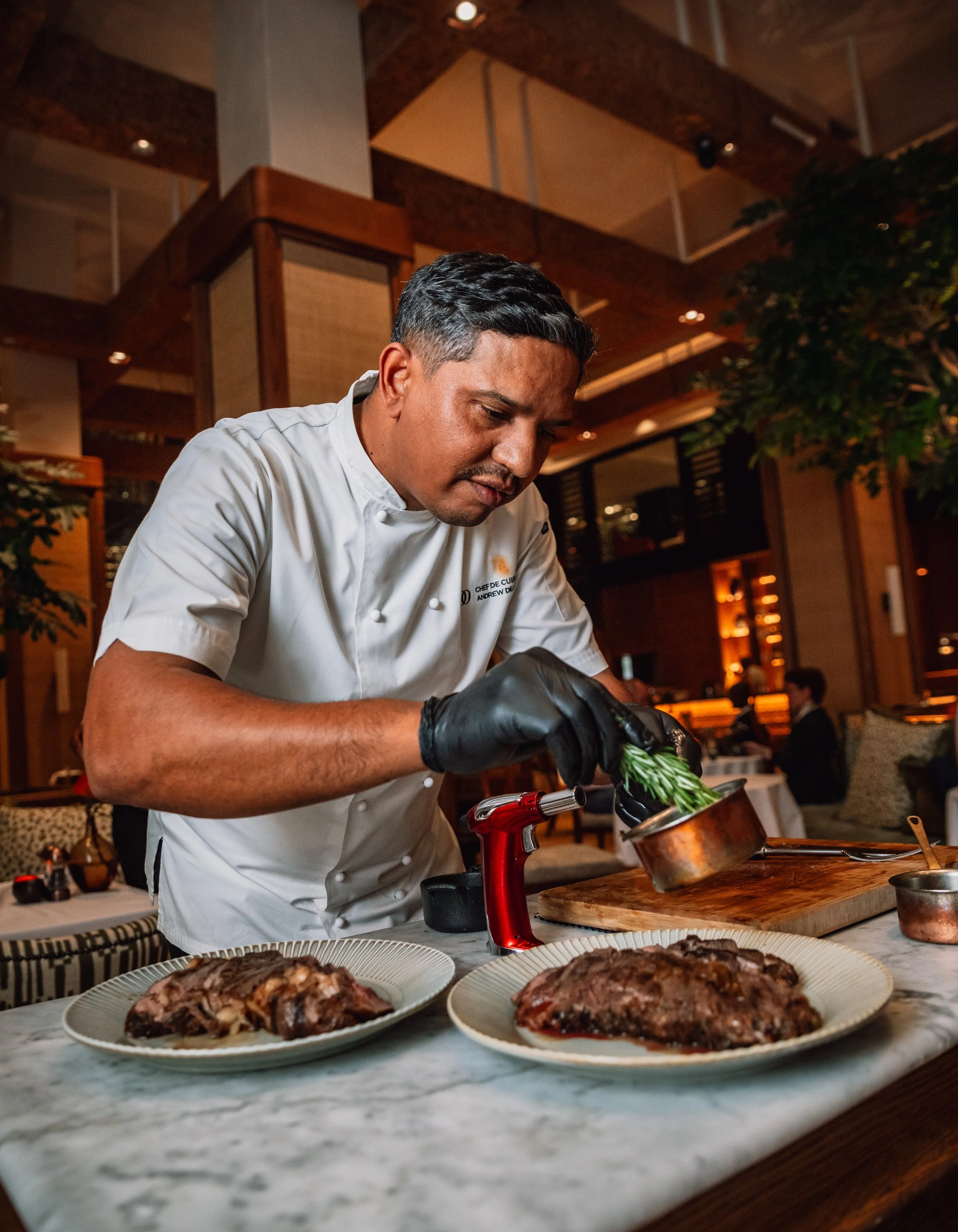 A chef preparing food in a restaurant kitchen, with plates of cooked meat on a marble countertop in the foreground.