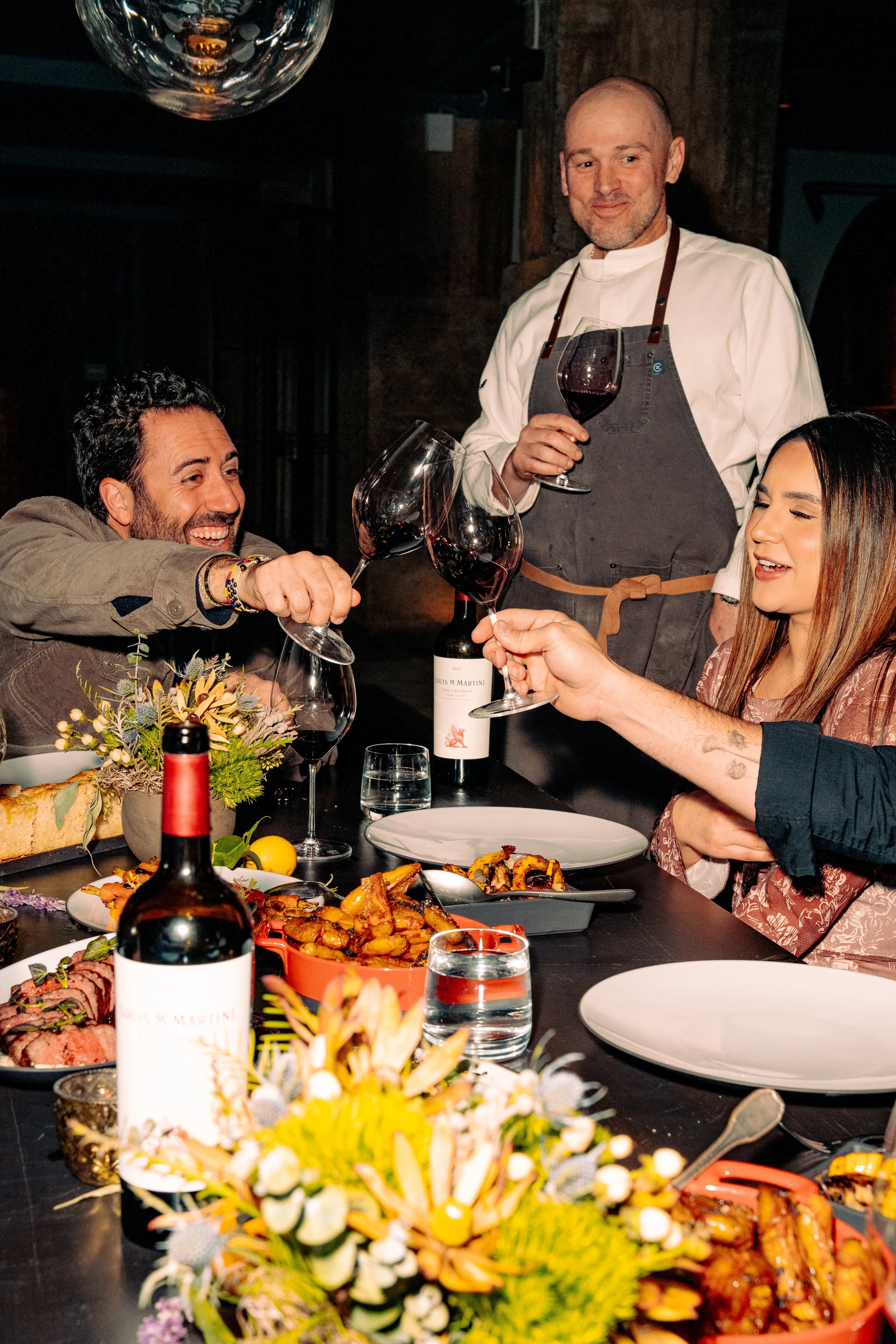 A group of people enjoying a dinner party with a chef serving wine, and a table with various dishes, a wine bottle, flowers, and utensils.