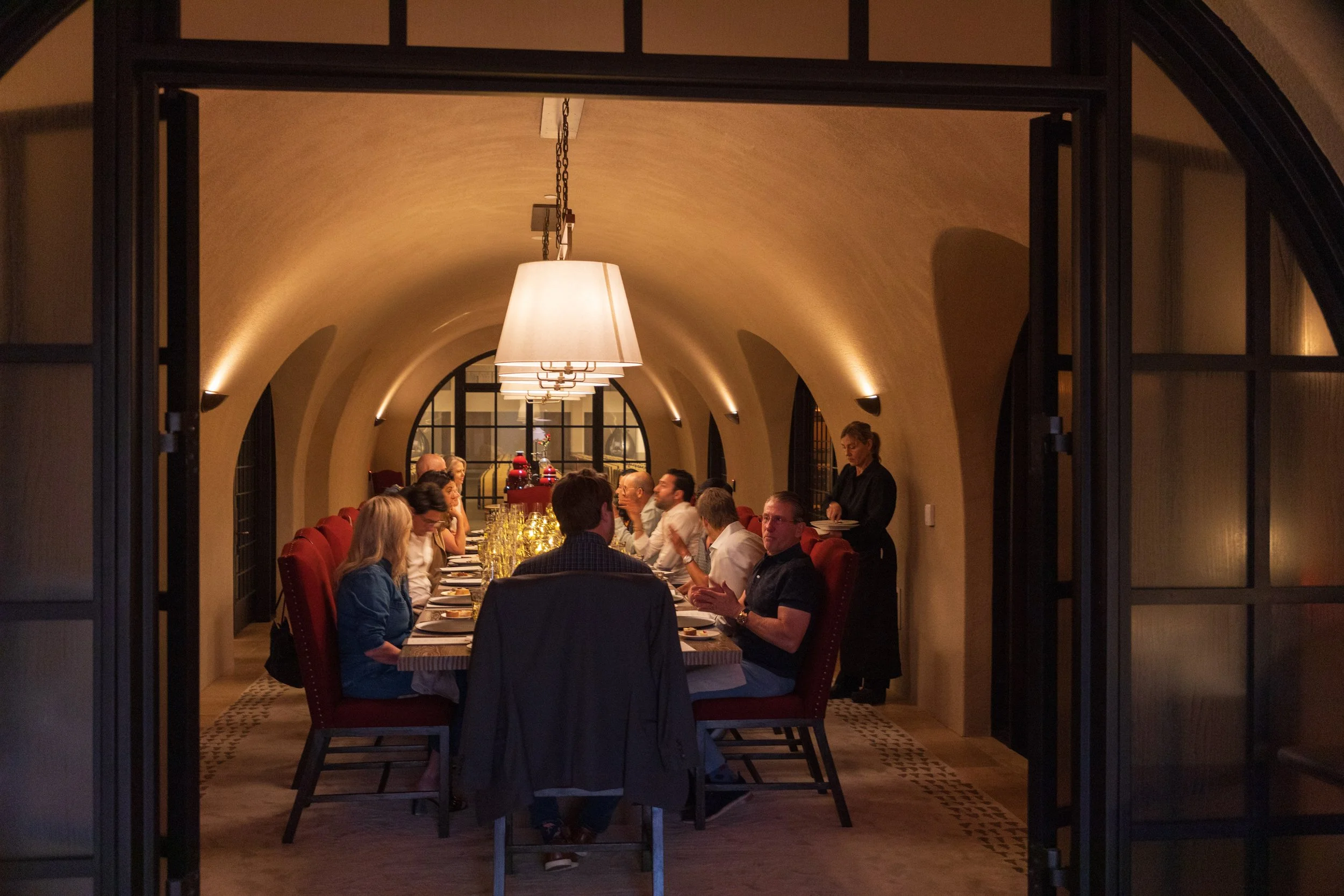 People dining at a long table in a warmly lit, arched-ceiling restaurant seen through a black metal door frame.