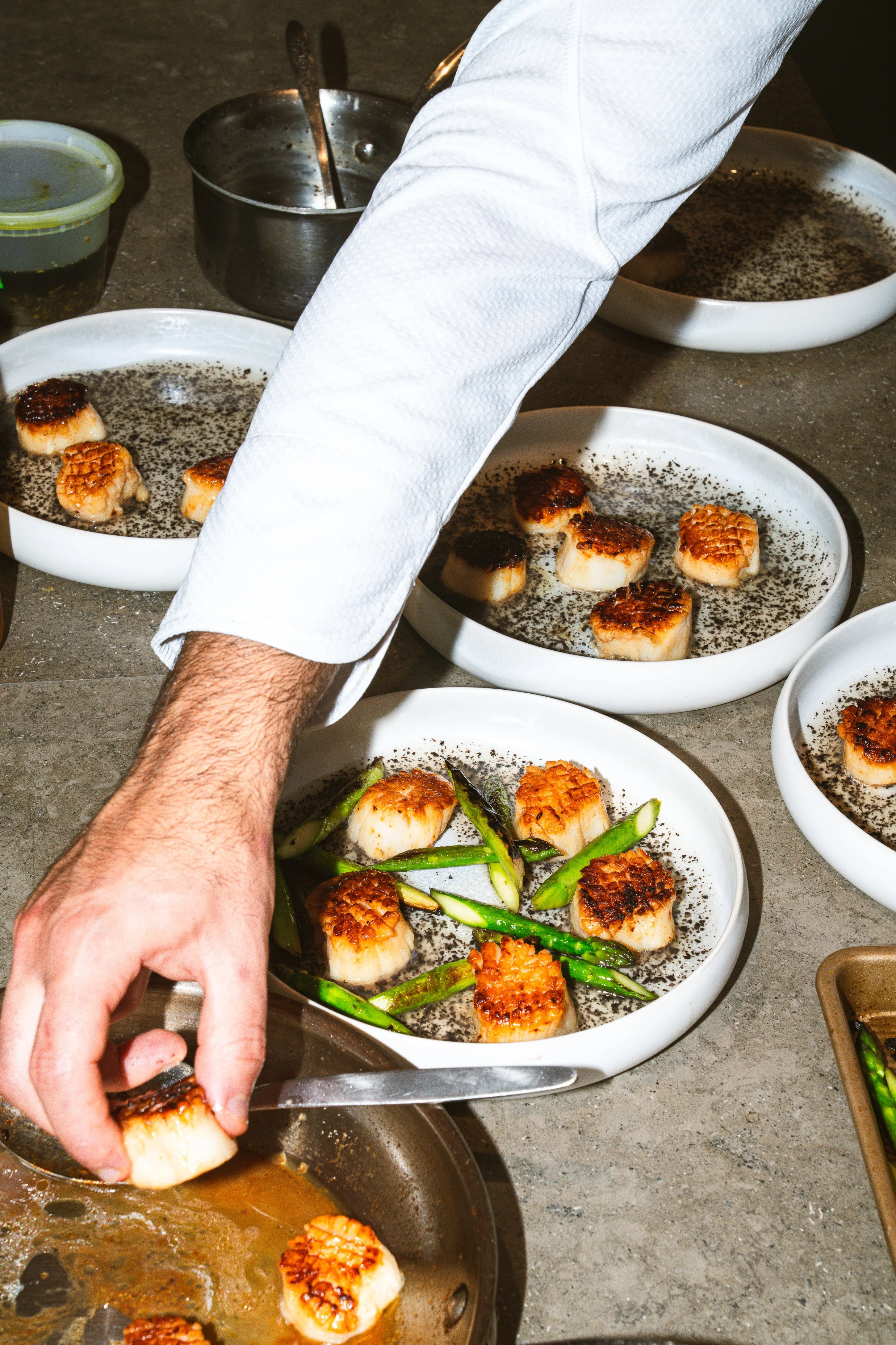Close-up of a chef's arm and hand preparing scallops with seared tops, in a kitchen setting, surrounded by bowls with ingredients and a pan with sauce.