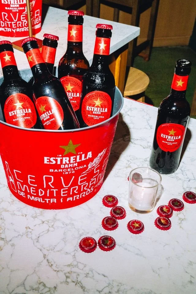 A red bucket filled with several bottles of Estrella Damm beer on a white marble table, with some loose bottle caps and an empty glass nearby.