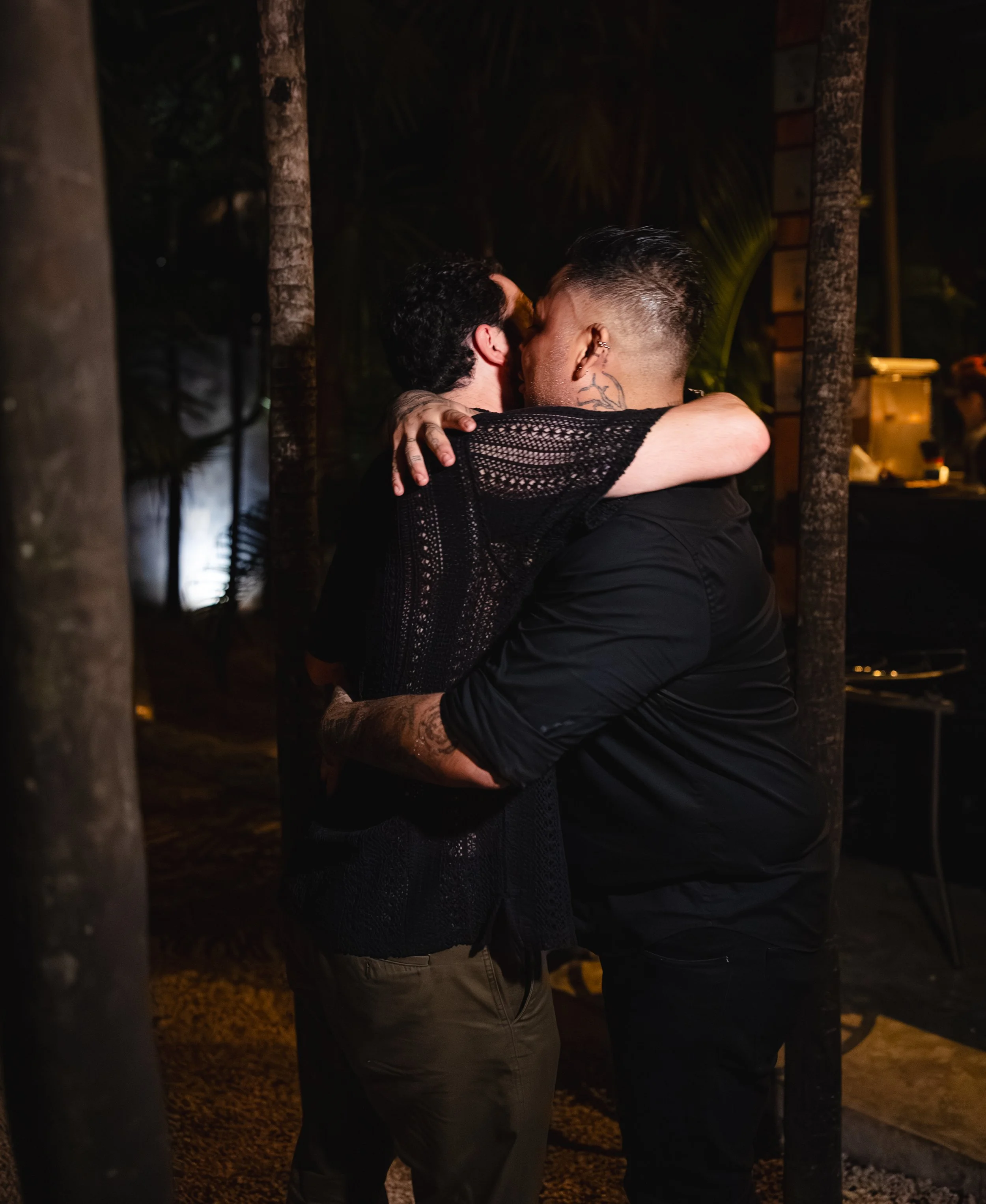 Two men embracing and kissing each other in a dimly lit outdoor setting at night, surrounded by trees and a wooden structure.