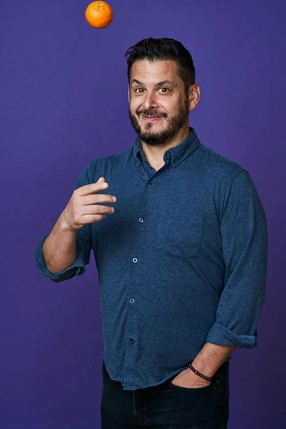 Man with a beard and short dark hair, wearing a blue button-up shirt with rolled-up sleeves, smiling and pointing to his chest, with an orange floating above his head, set against a purple background.