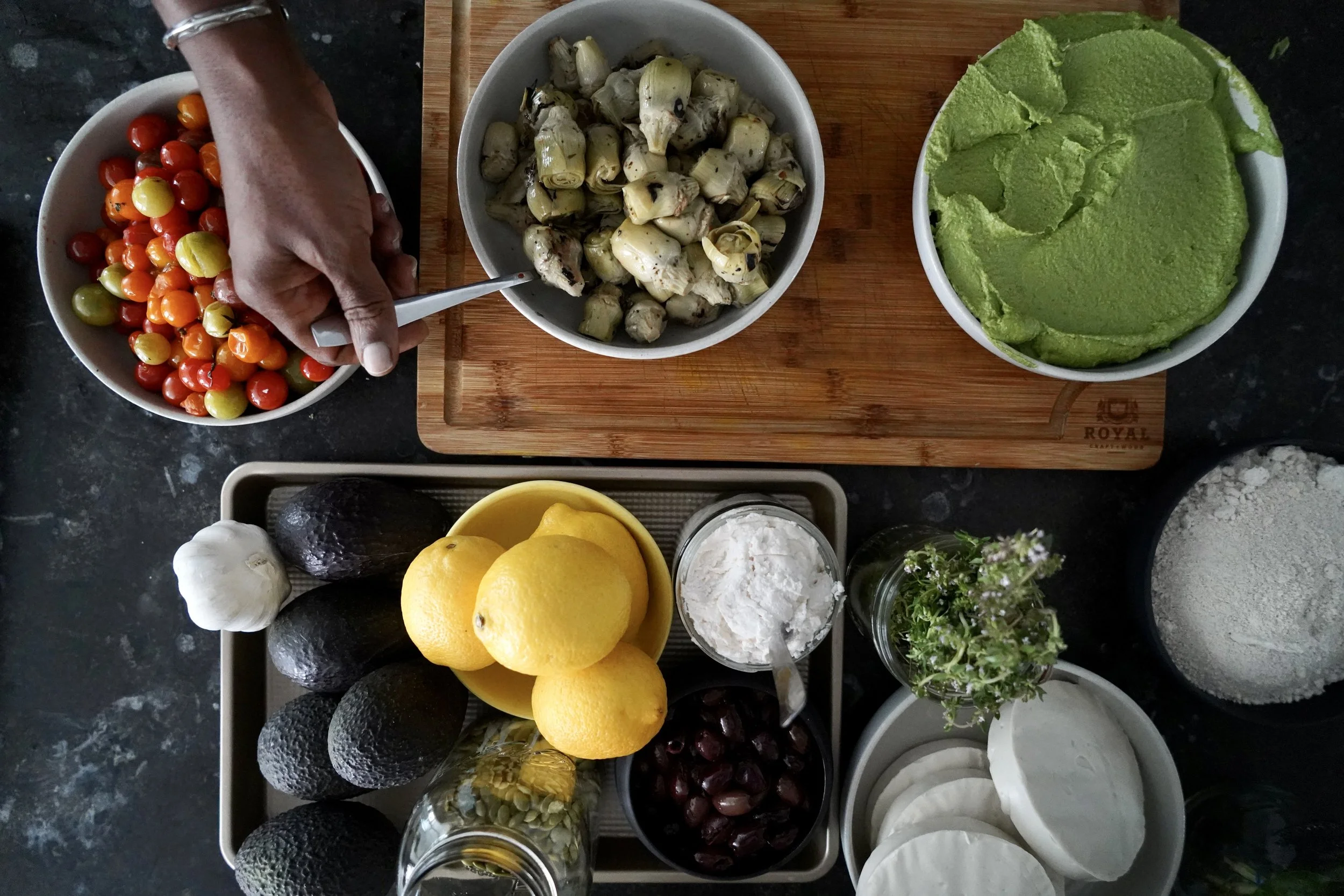 A variety of fresh fruits and vegetables arranged on a black countertop. There are cherry tomatoes in a bowl, artichoke hearts in a bowl, avocados, lemons, garlic, condiments, grapes, and herbs. A hand is reaching into the cherry tomatoes.