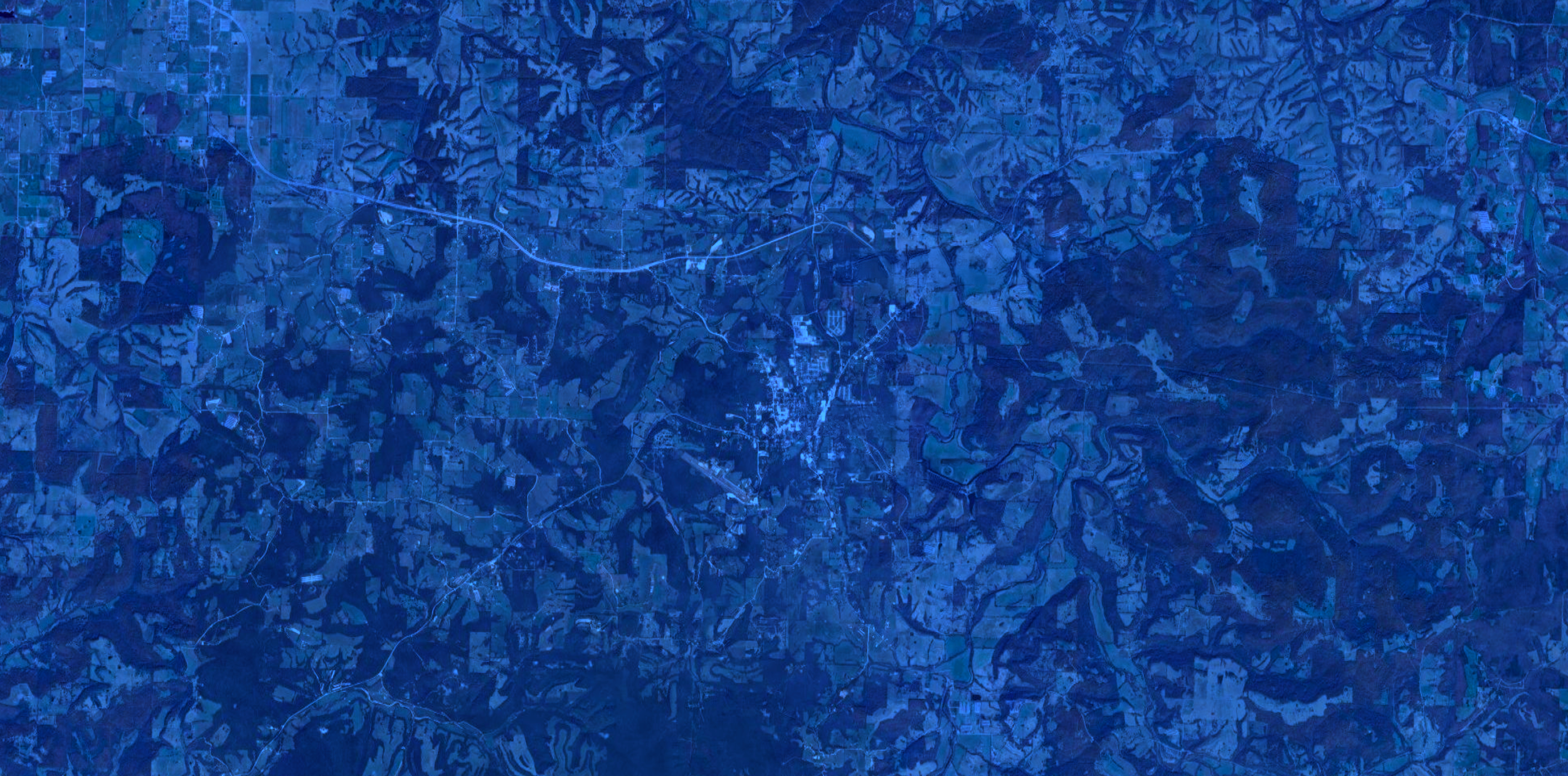 Satellite image showing a rural landscape with winding roads, small clusters of buildings, and patches of farmland in shades of blue.