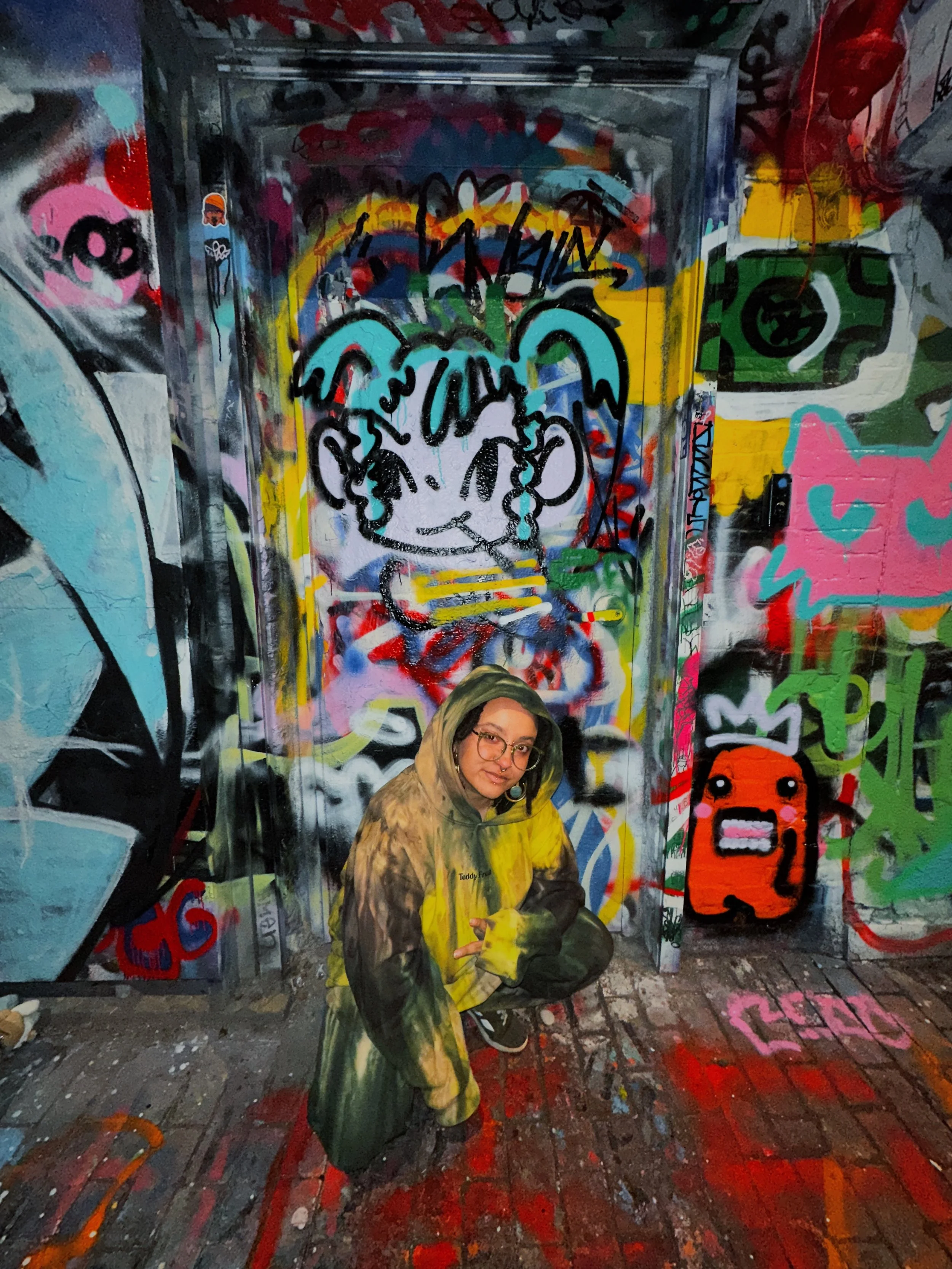 Kai in a camouflage-ish patterned outfit and glasses crouches in front of a wall and door covered in colorful graffiti art, inside the legal wall alley in Cambridge also decorated with graffiti on the ground and walls.