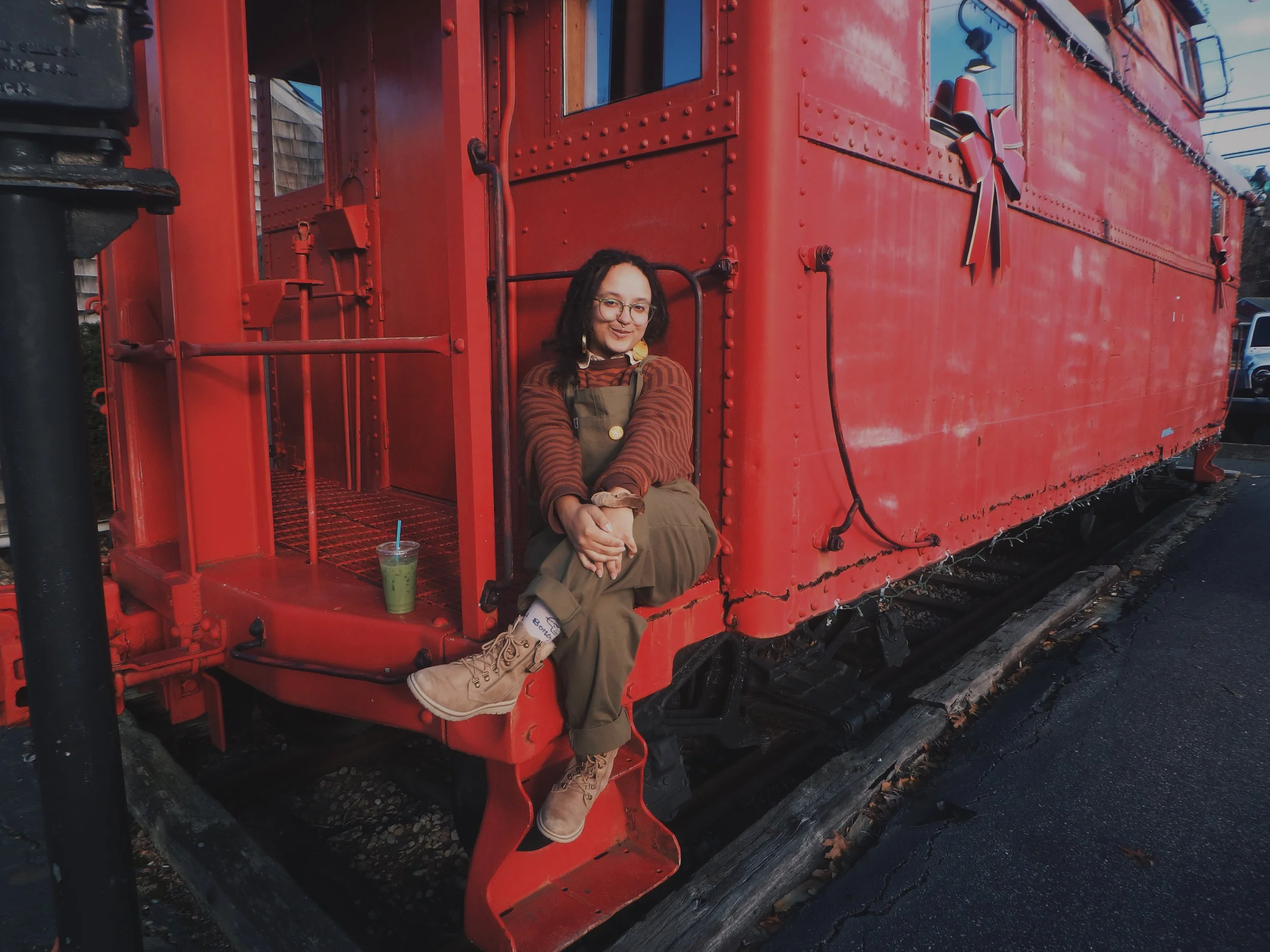 A person with glasses, locced hair, and a striped shirt sitting on the steps of a red train carriage, holding their knees with a green drink placed beside them.