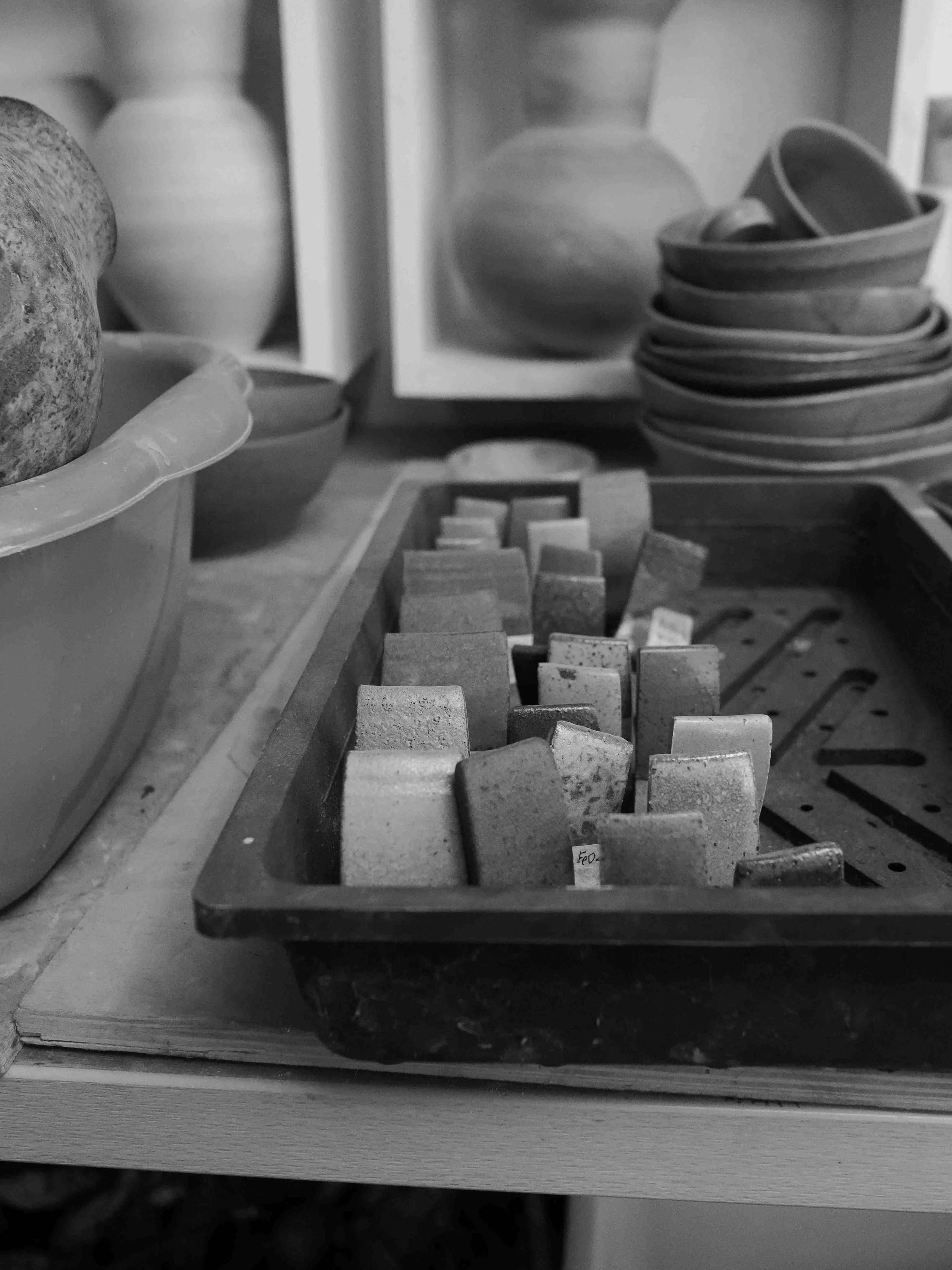 A black and white photo of a ceramics studio with various pottery pieces, including rectangular blocks in a tray, bowls stacked to the right, and vases in the background.