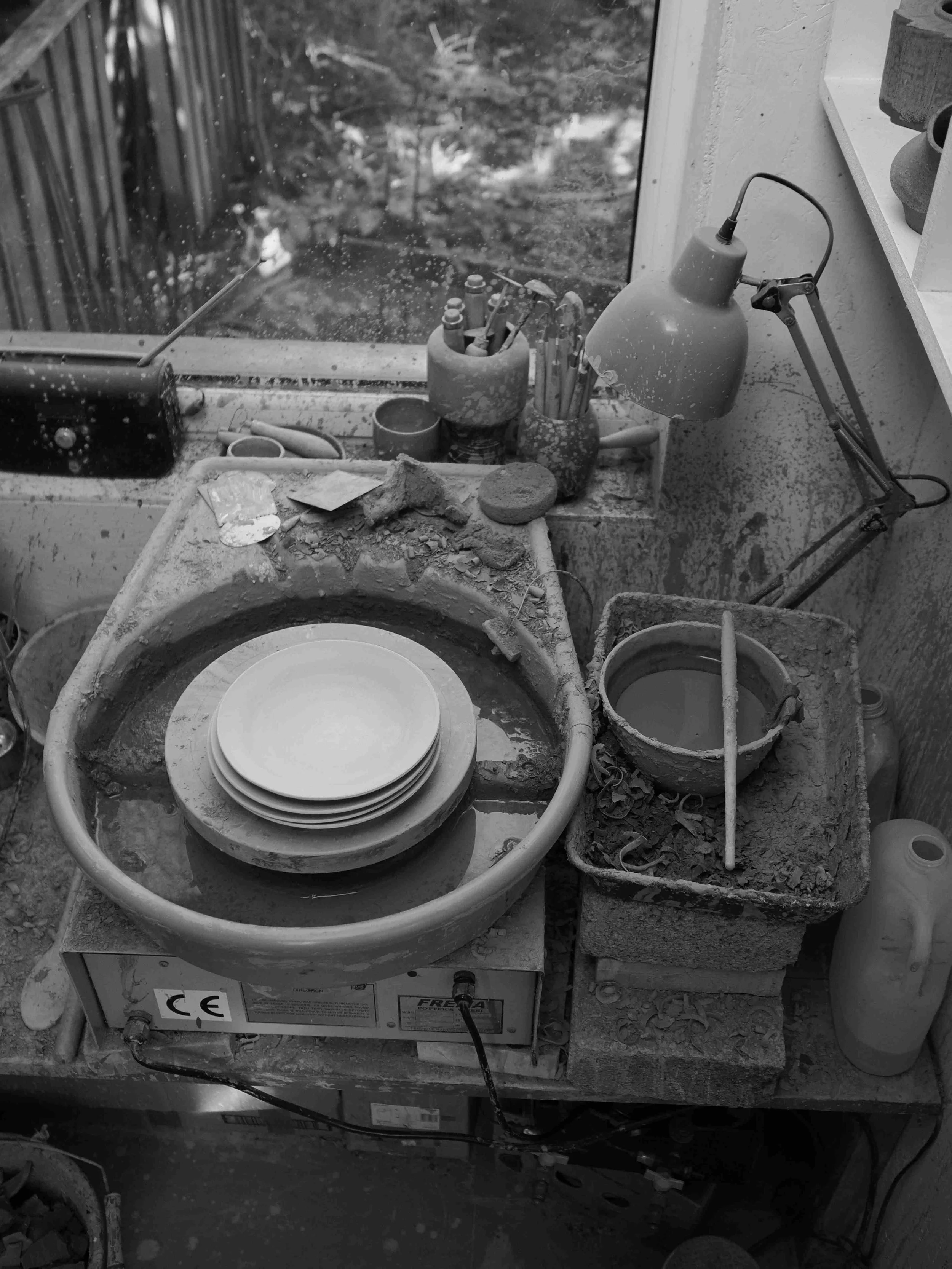 A pottery wheel in a studio surrounded by pottery tools and supplies, next to a window showing a garden.