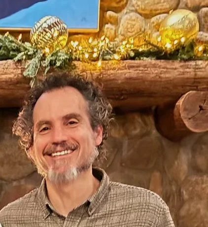A man with curly hair and a beard smiling in front of a decorated fireplace with Christmas ornaments and greenery.