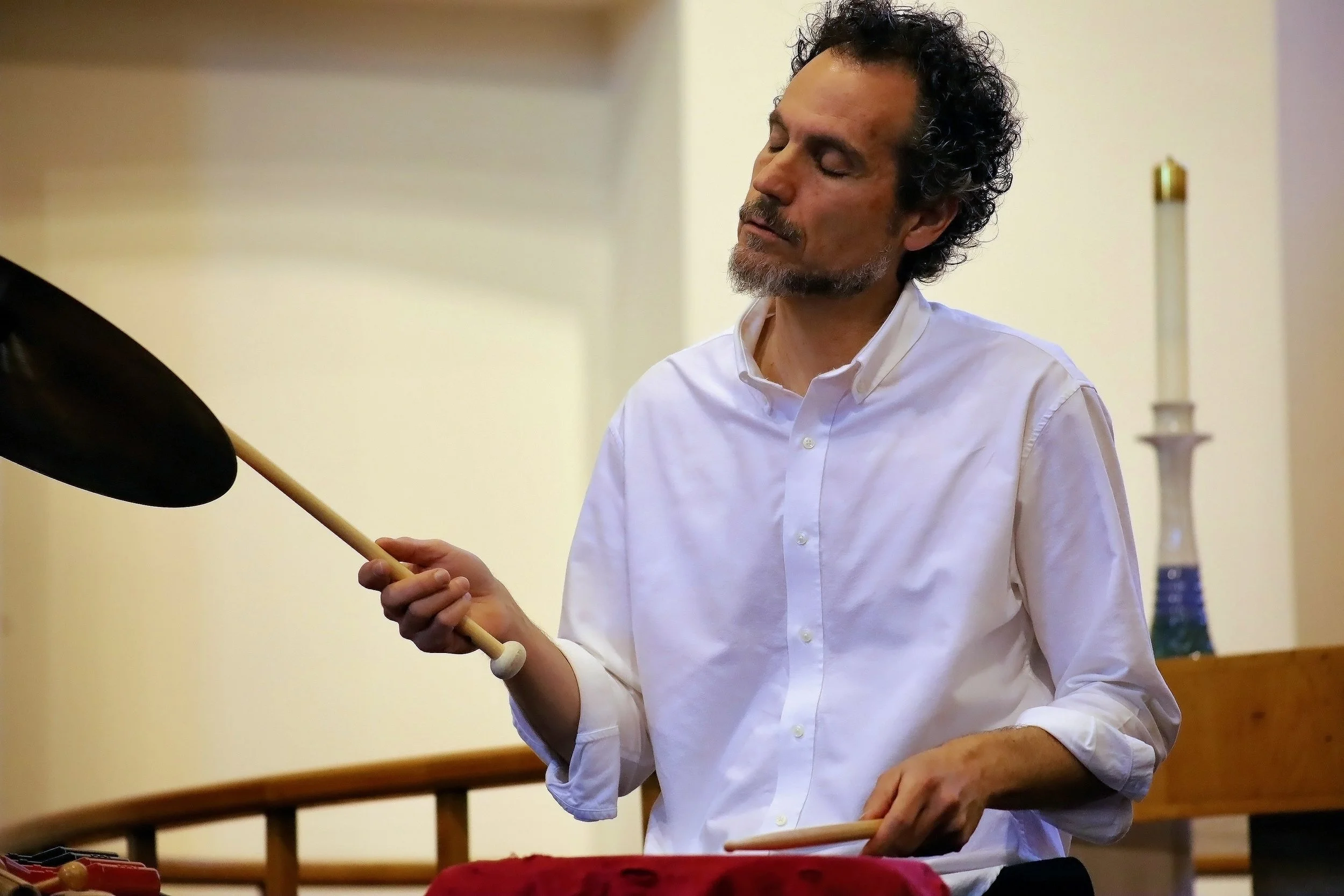 A man with curly black hair and a beard, wearing a white dress shirt, is playing a cymbal with a drumstick in an indoor setting. There is a candle and a glass vase in the background.