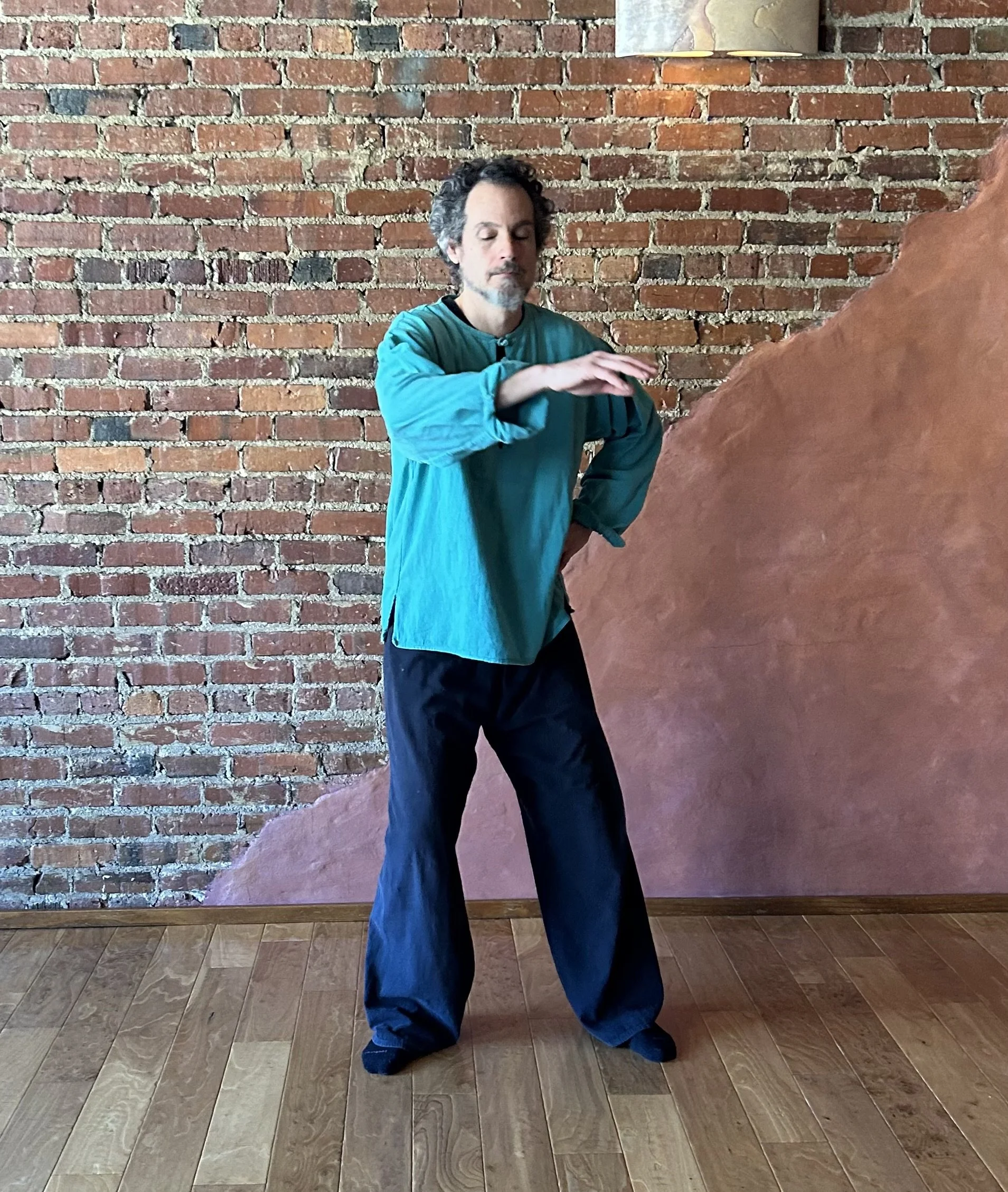 A man with curly hair and a beard practicing Tai Chi indoors, standing with one arm extended and the other hand on his hip in front of a brick wall.