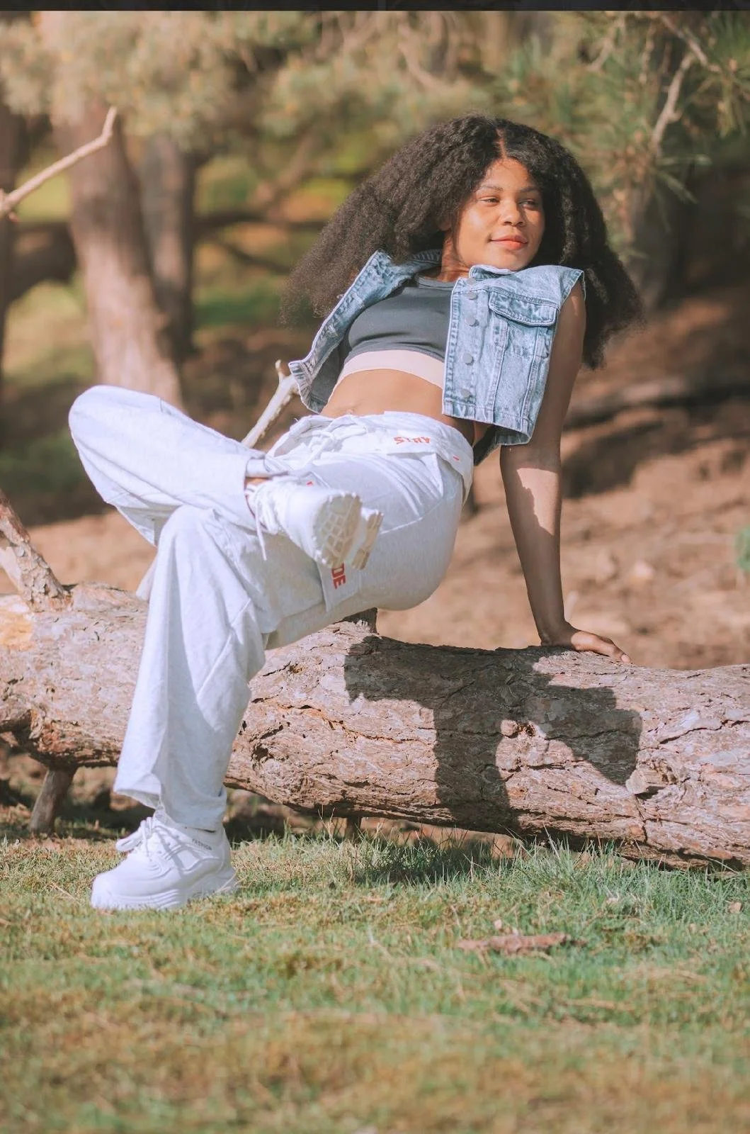 A woman with natural curly hair sits on a fallen tree trunk outdoors, basking in the sunlight, with a blurred background of trees and grass.