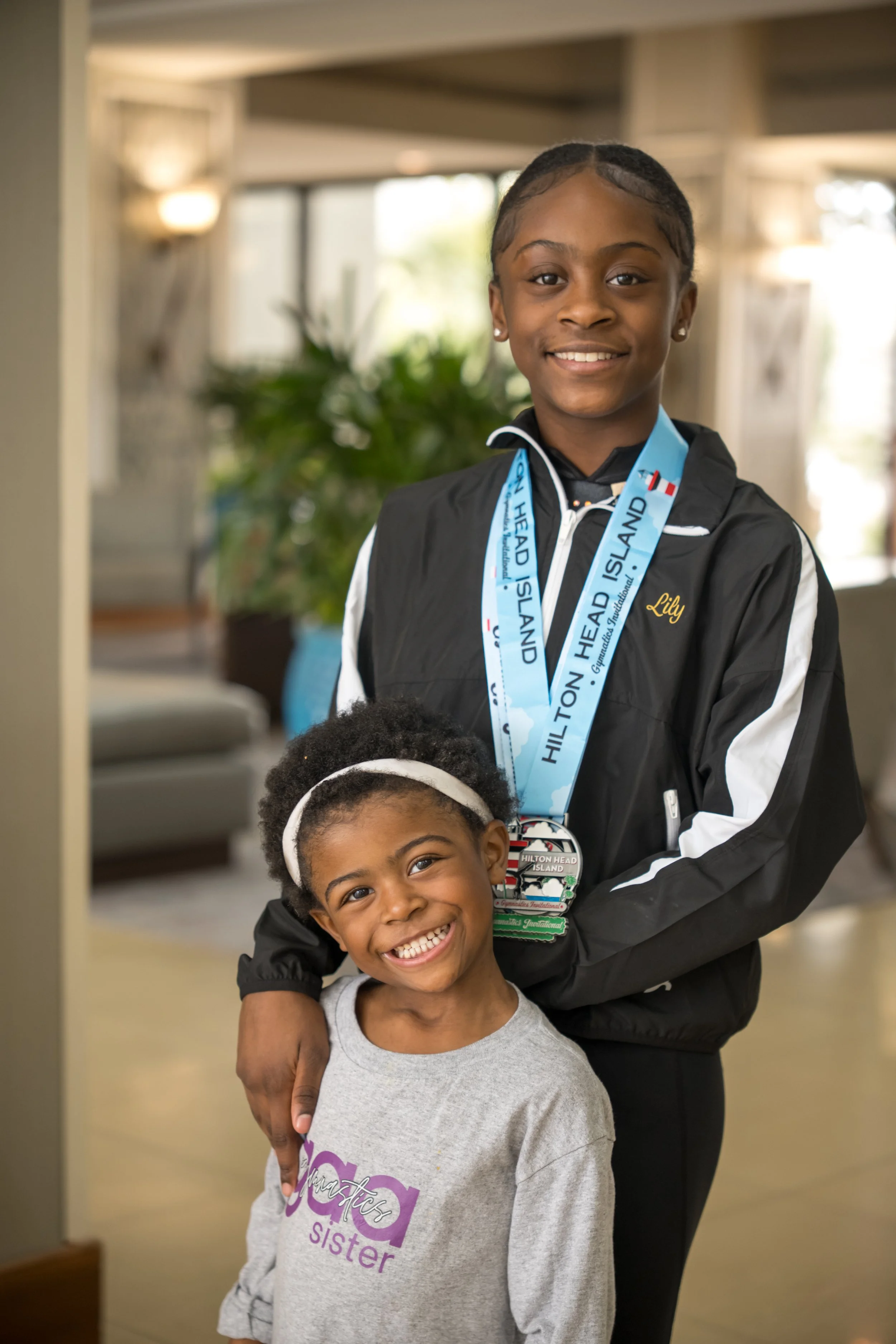 Two smiling girls indoors, one older girl wearing a black jacket with a medal around her neck and the other younger girl wearing a gray t-shirt. The older girl has braids and the younger girl has curly hair with a headband.