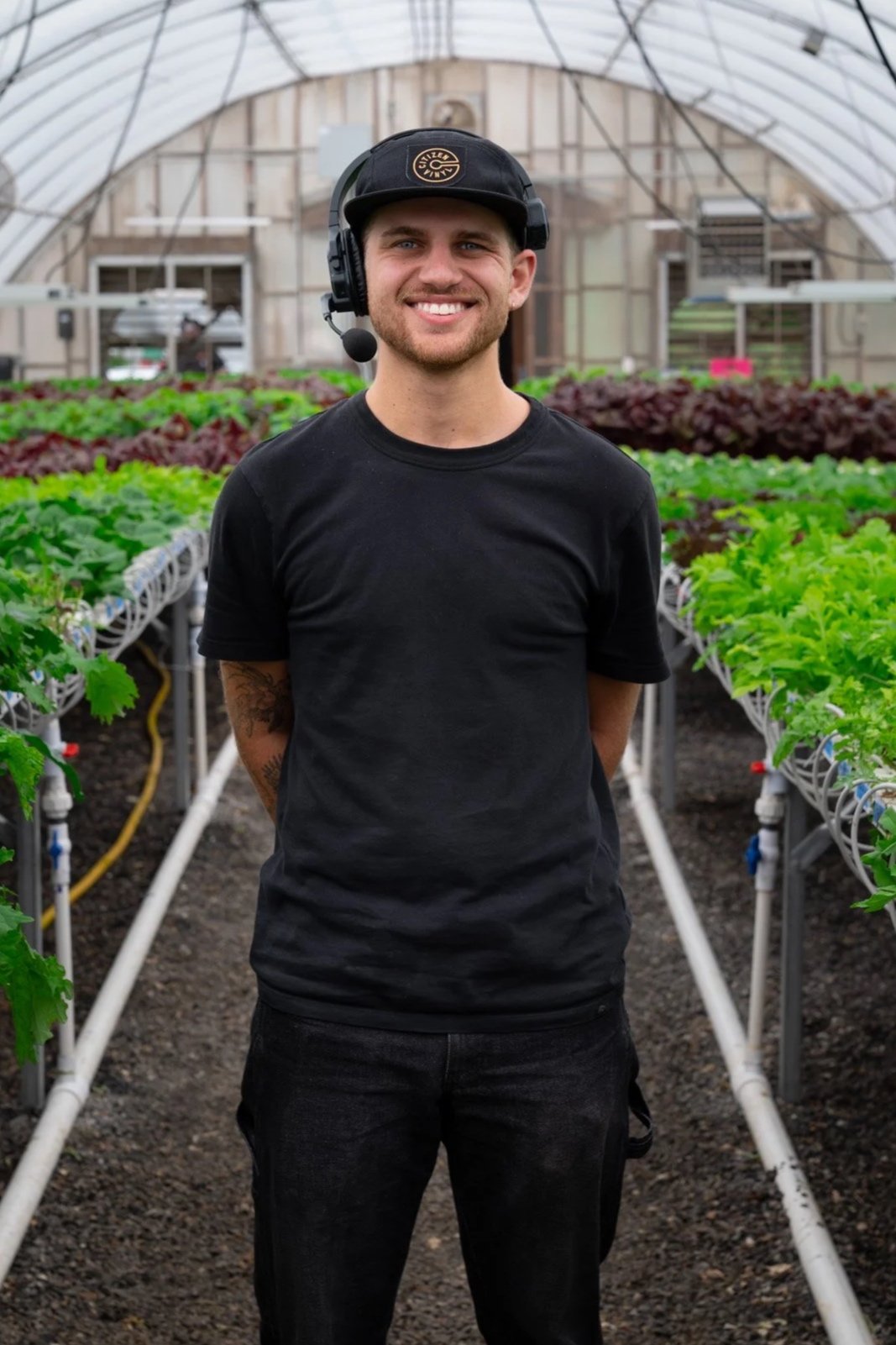 A young man with a headset and microphone, wearing a black T-shirt and black pants, standing inside a greenhouse surrounded by green and purple lettuce plants.