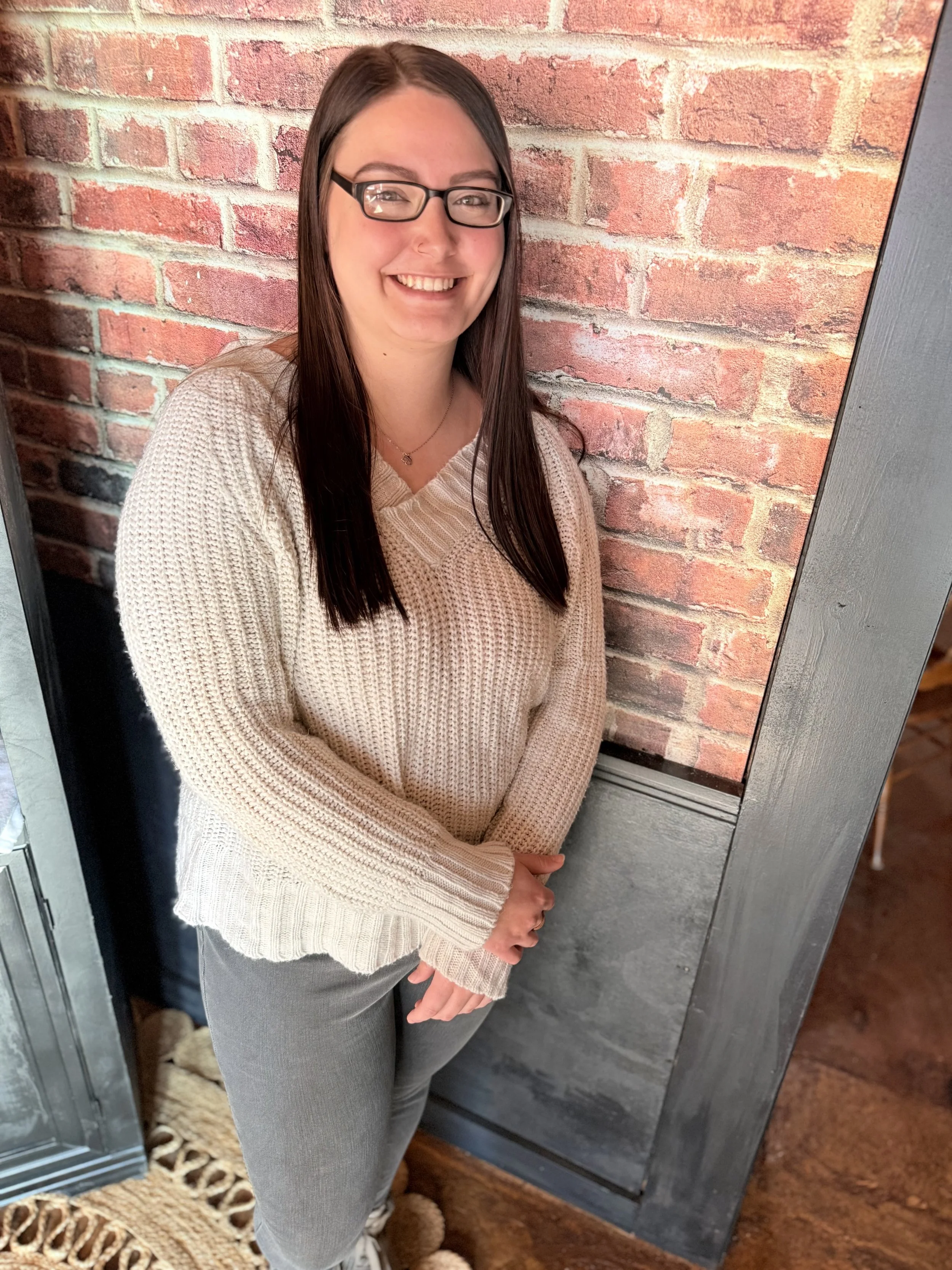 A woman with long dark hair, glasses, and a smile, wearing a pink top and black pants, sitting on a teal sofa with a colorful decorative pillow, in a living room with abstract wall art in the background.