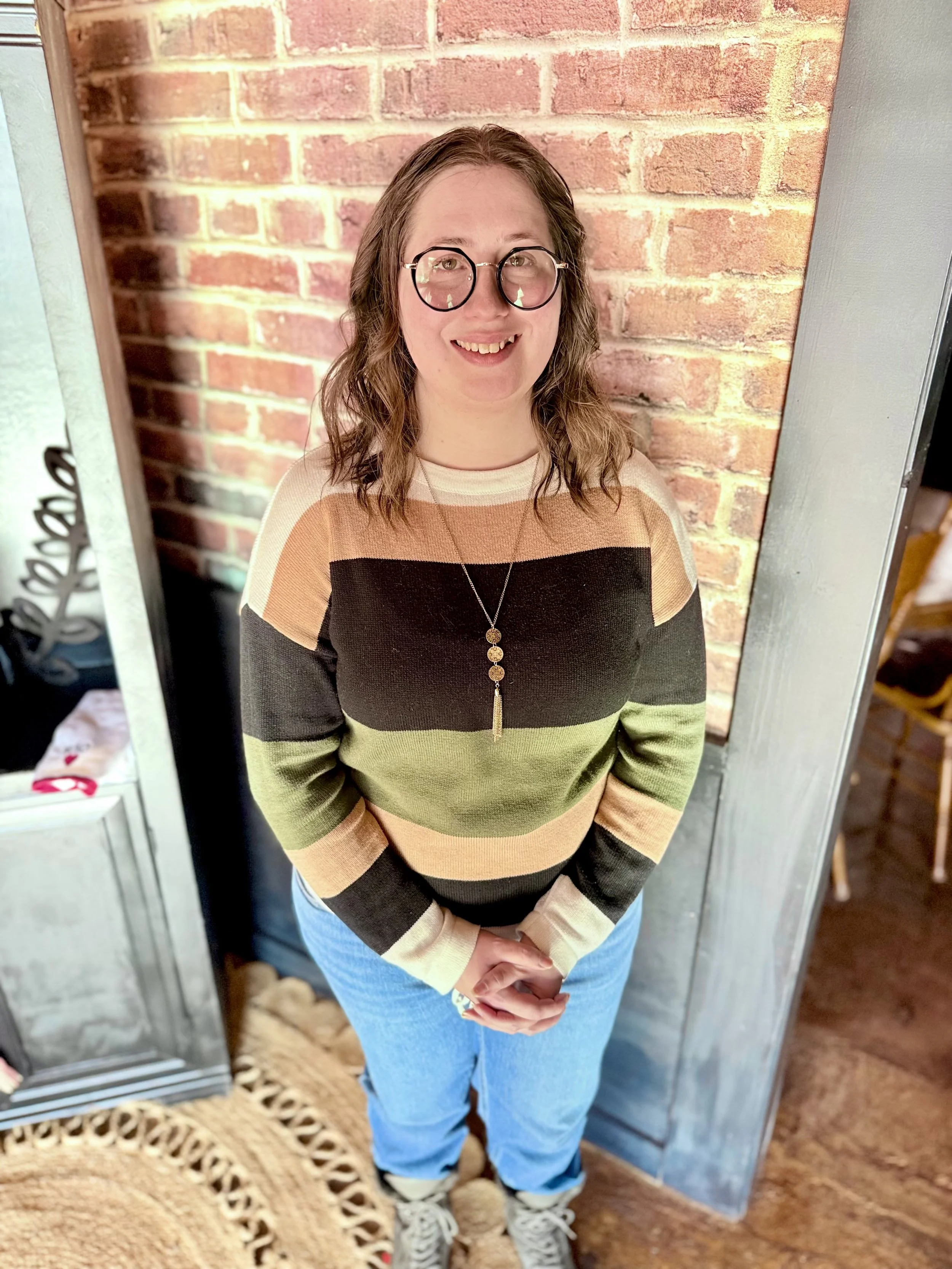 A woman with glasses and long brown hair, smiling and wearing a blue dress with a pink and green floral pattern, standing outdoors in a parking lot with trees and grass in the background.