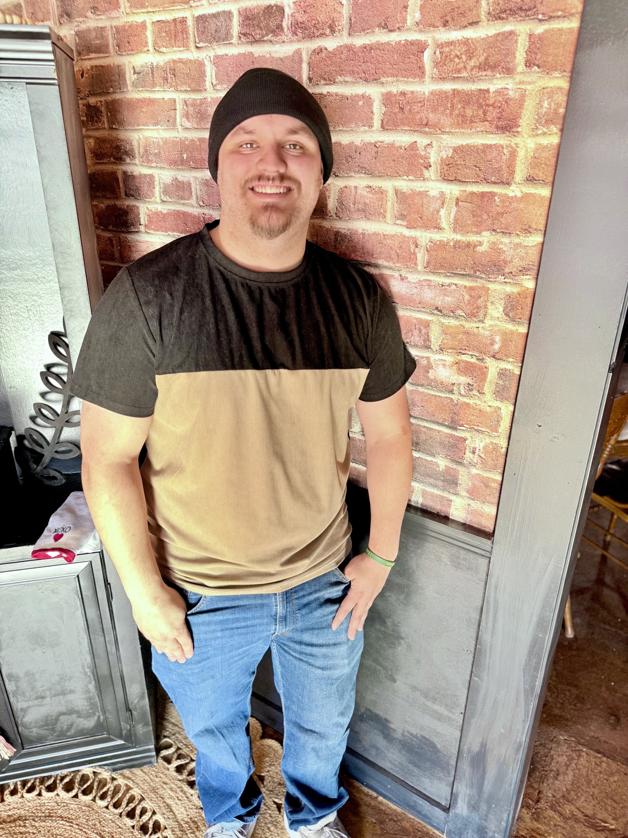 A smiling man wearing a blue short-sleeve shirt, black beanie, and gold chain outdoors with trees in the background.