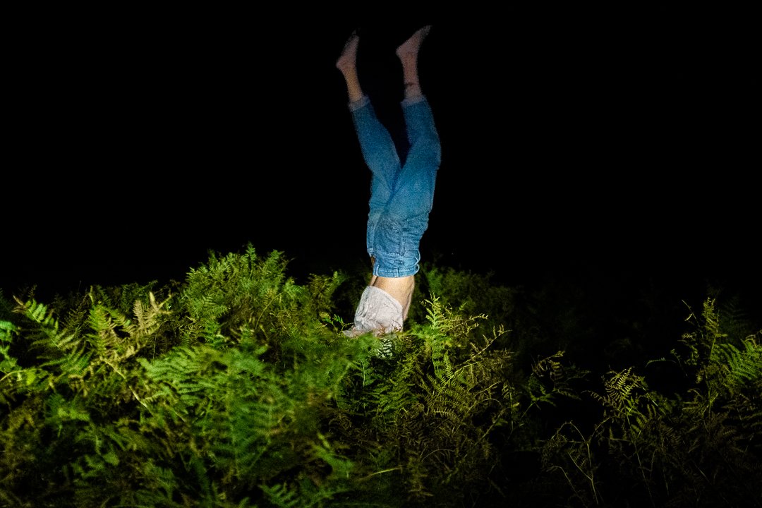 Eine Person steht im Dunkeln auf den Händen, umgeben von Farnen. Die Beine sind in blauen Jeans gekleidet.