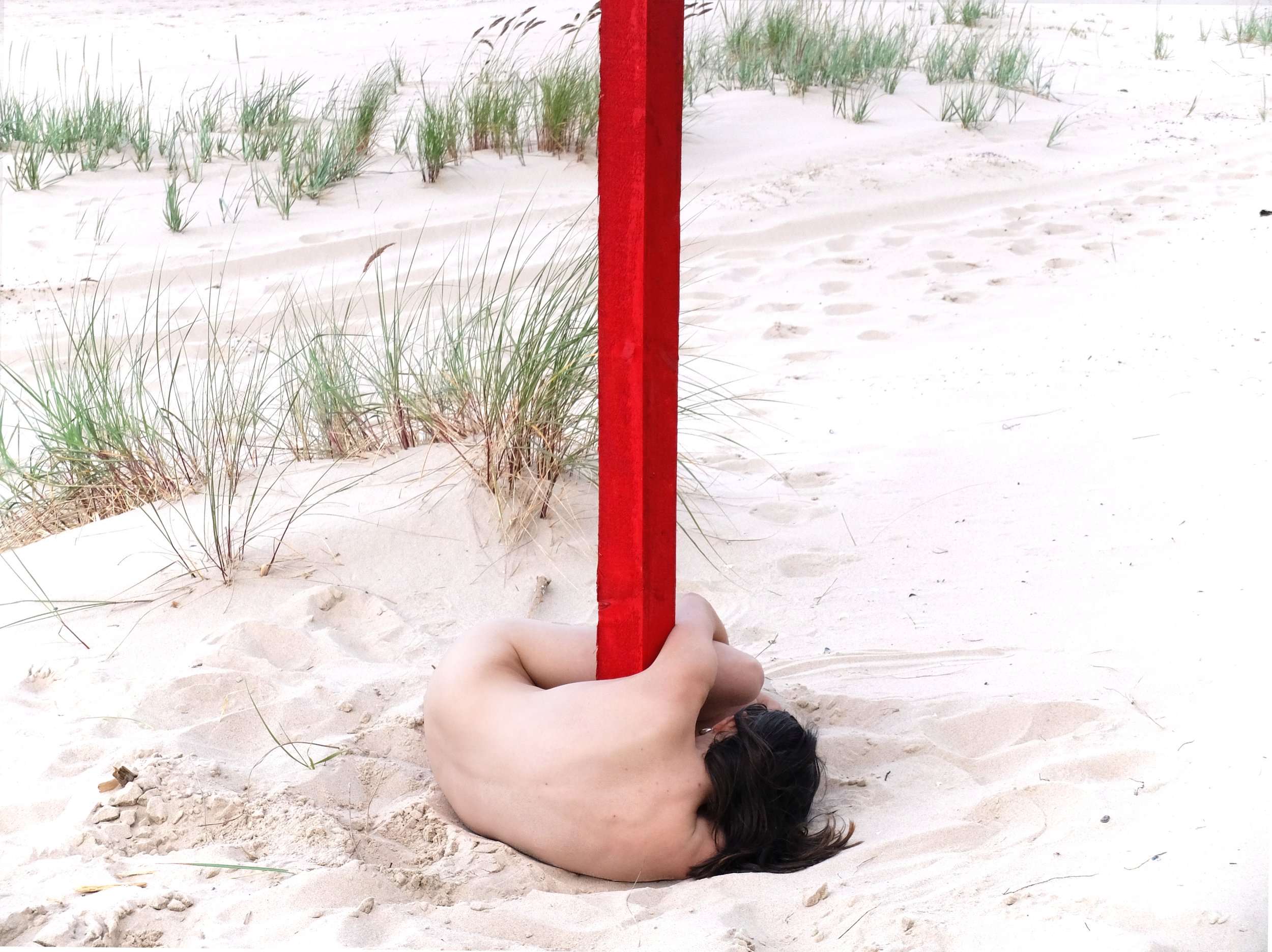 Eine nackte Person umarmt einen roten Pfahl im Sand, umgeben von Gräsern.
