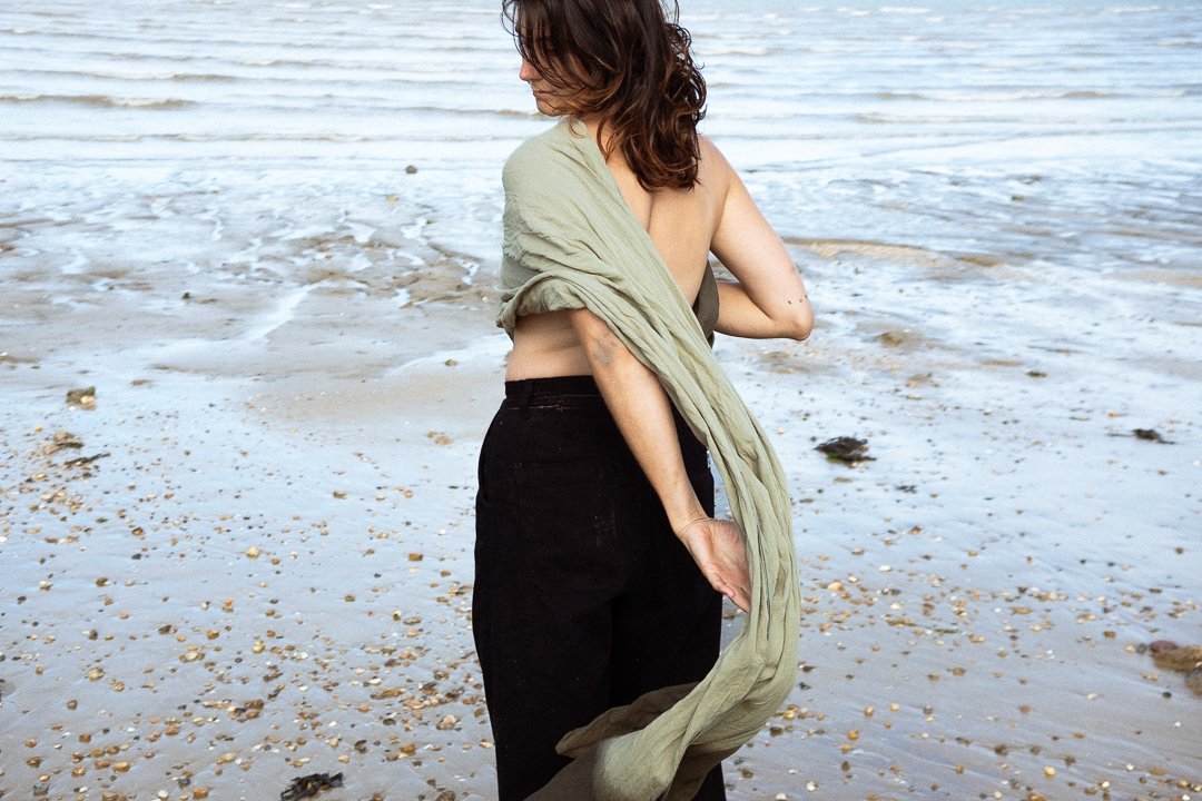 Frau steht am Strand, trägt schwarzes Hosen und ein grünes Tuch. Sie blickt aufs Meer.