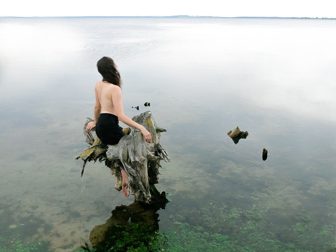 Person sitzt oberkörperfrei auf einem Baumstumpf im Wasser, blickt aufs Meer hinaus.