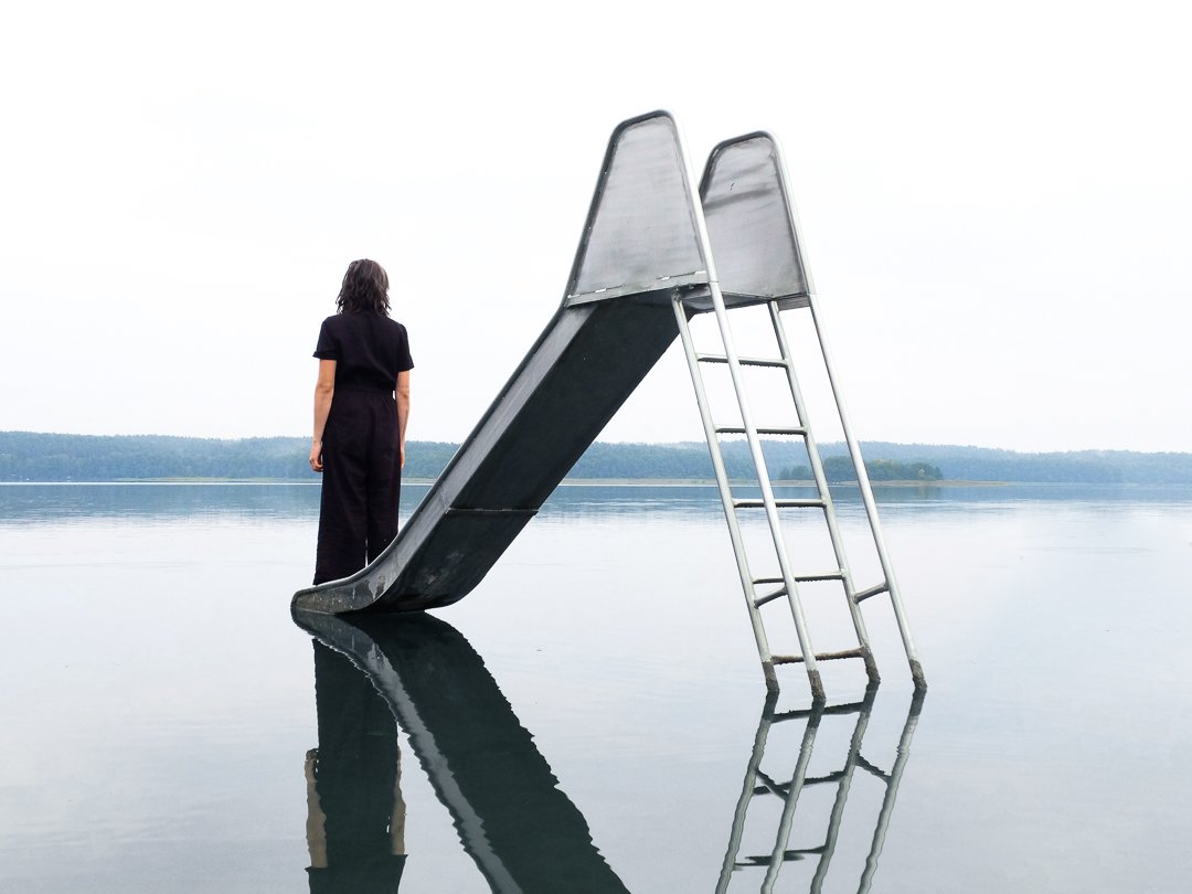Person in schwarzer Kleidung steht am Ende einer Rutsche, die auf einer Wasseroberfläche reflektiert wird.