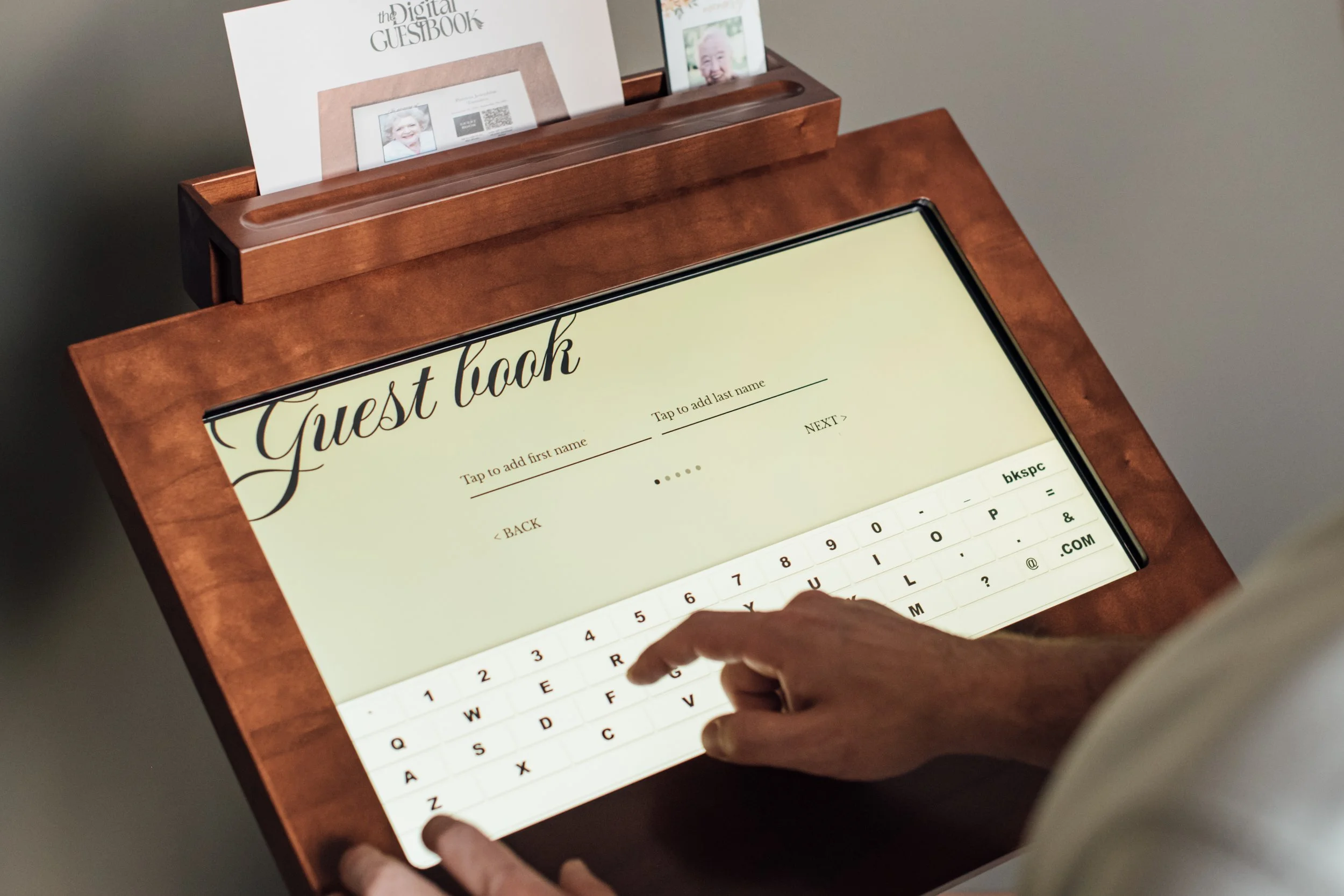 A person is using a touchscreen digital guestbook framed with a wooden border. The screen displays a welcome message and a virtual keyboard for typing. There are printed signs and photos at the top of the frame, with one sign reading 'The Digital Guestbook'.