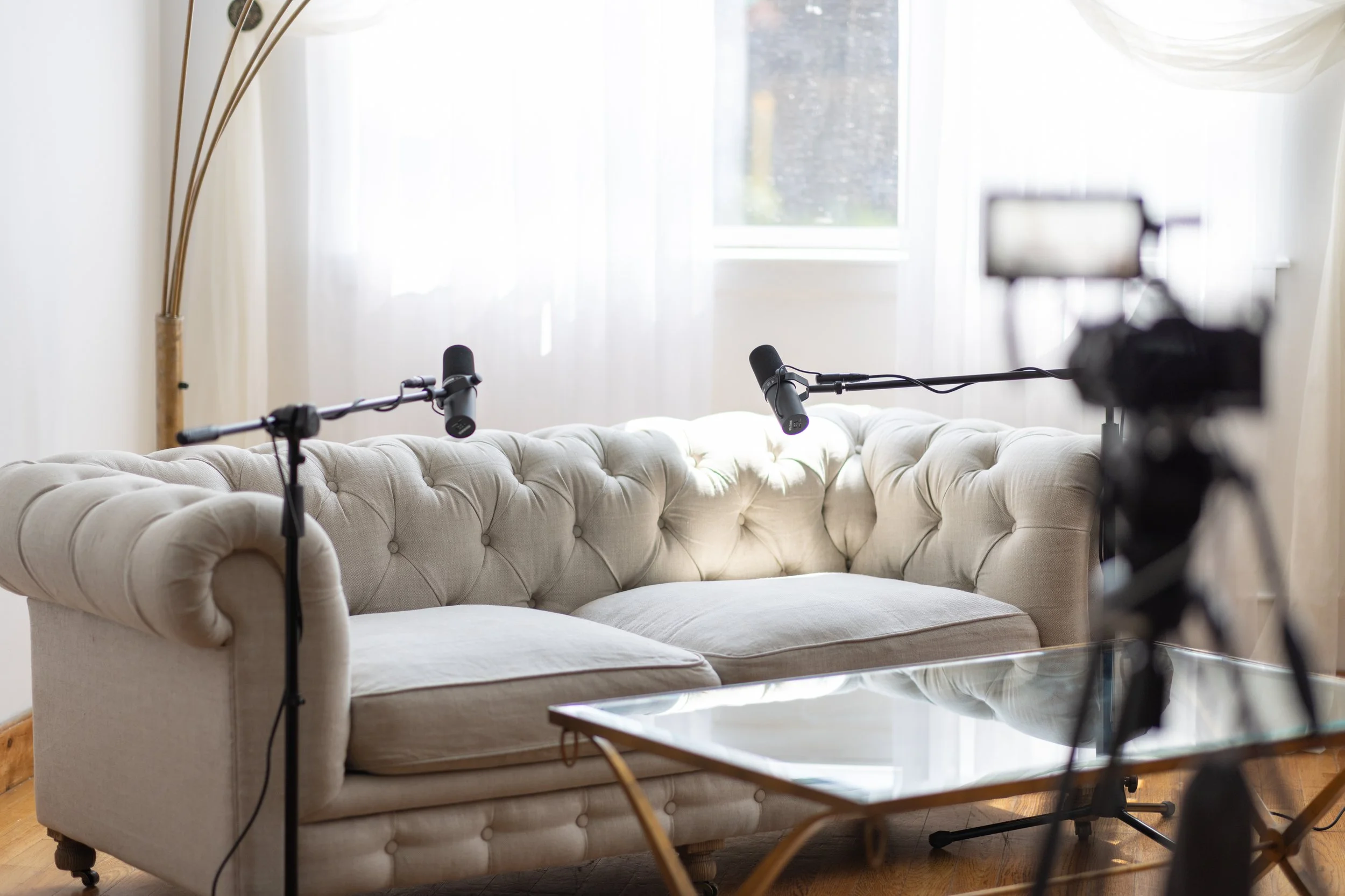 A cream-colored tufted sofa in front of a window with white curtains, with two microphones on stands and a camera on a tripod set up for recording or streaming in a well-lit room.
