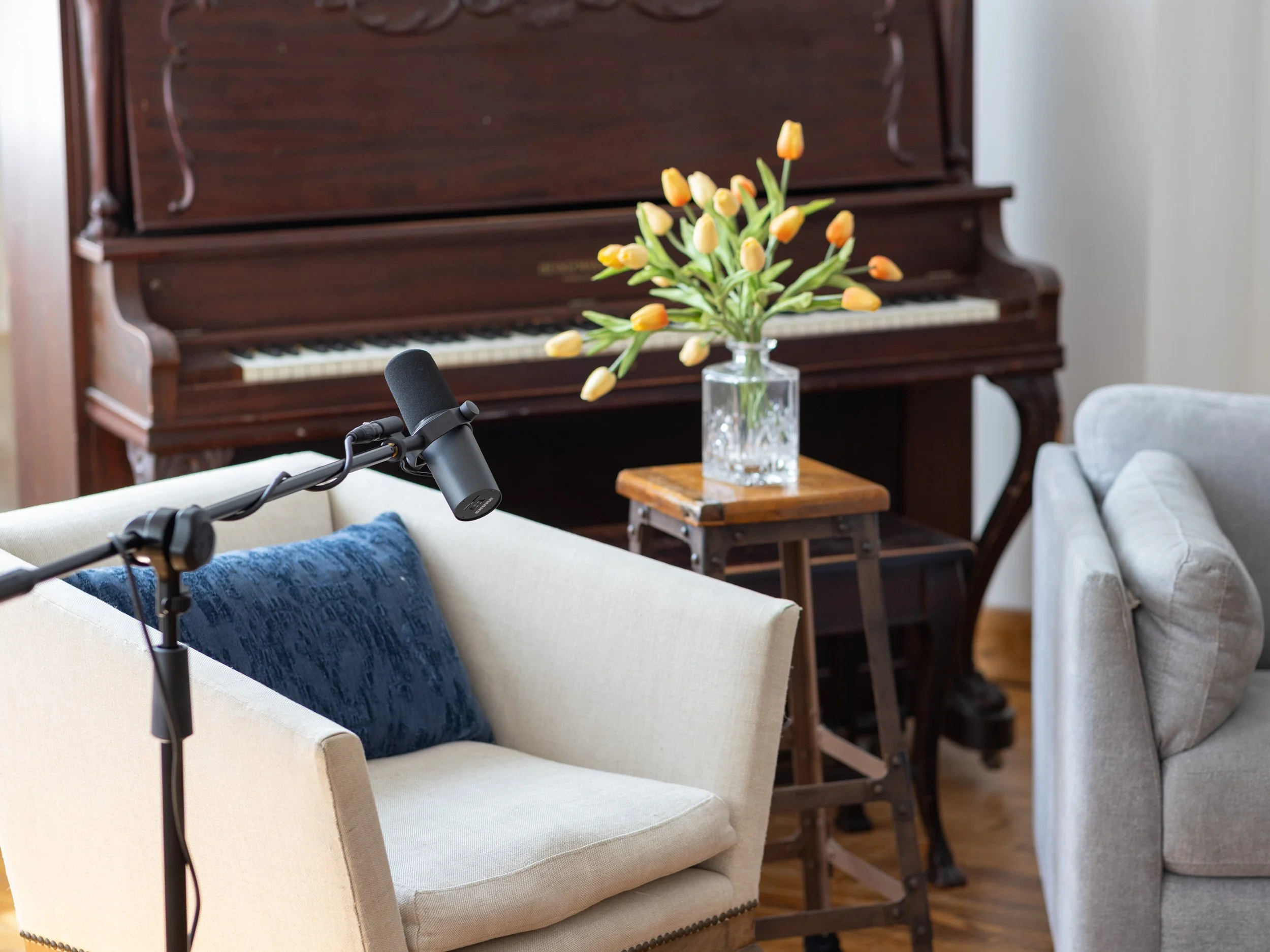 Interior room with a vintage wooden piano, a glass vase with yellow tulips on a small wooden table, a beige armchair with a blue cushion, a gray sofa with a matching pillow, and a professional microphone on a stand.