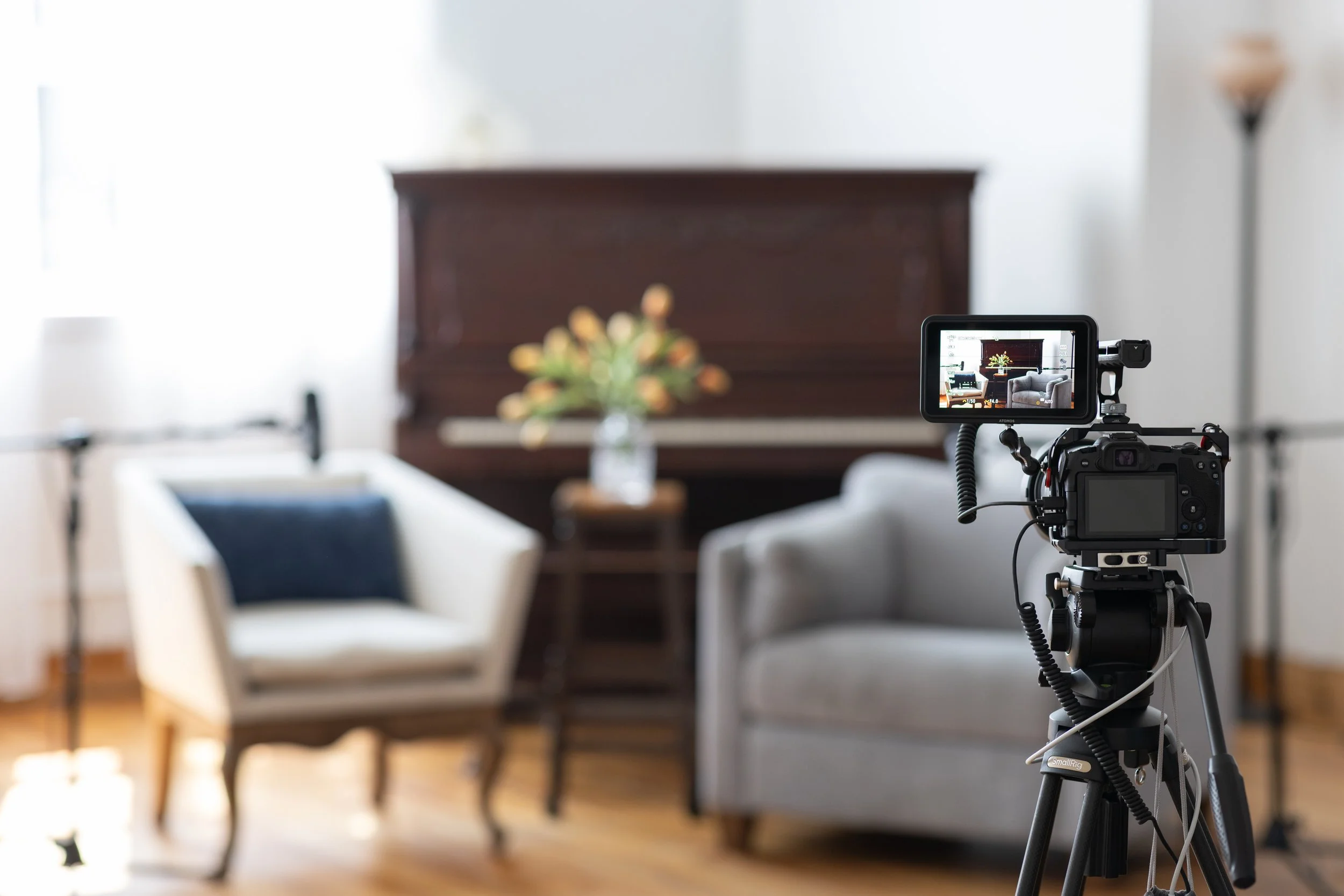 A camera on a tripod recording a living room setup with a piano, armchair, sofa, and a side table with a flower vase.
