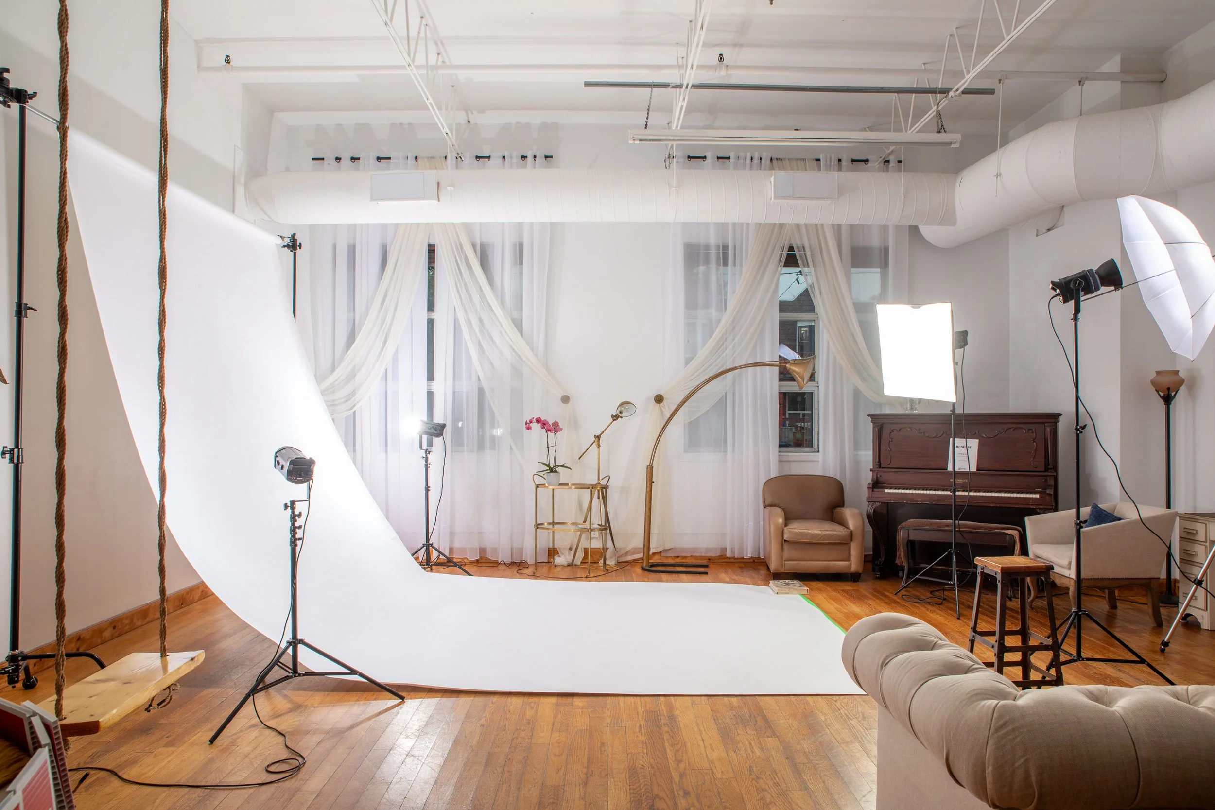 Indoor photography studio with a white backdrop, professional lighting equipment, sheer curtains on windows, a small table with a pink orchid, a brown armchair, a vintage brown piano, a beige armchair, and a gray tufted sofa.